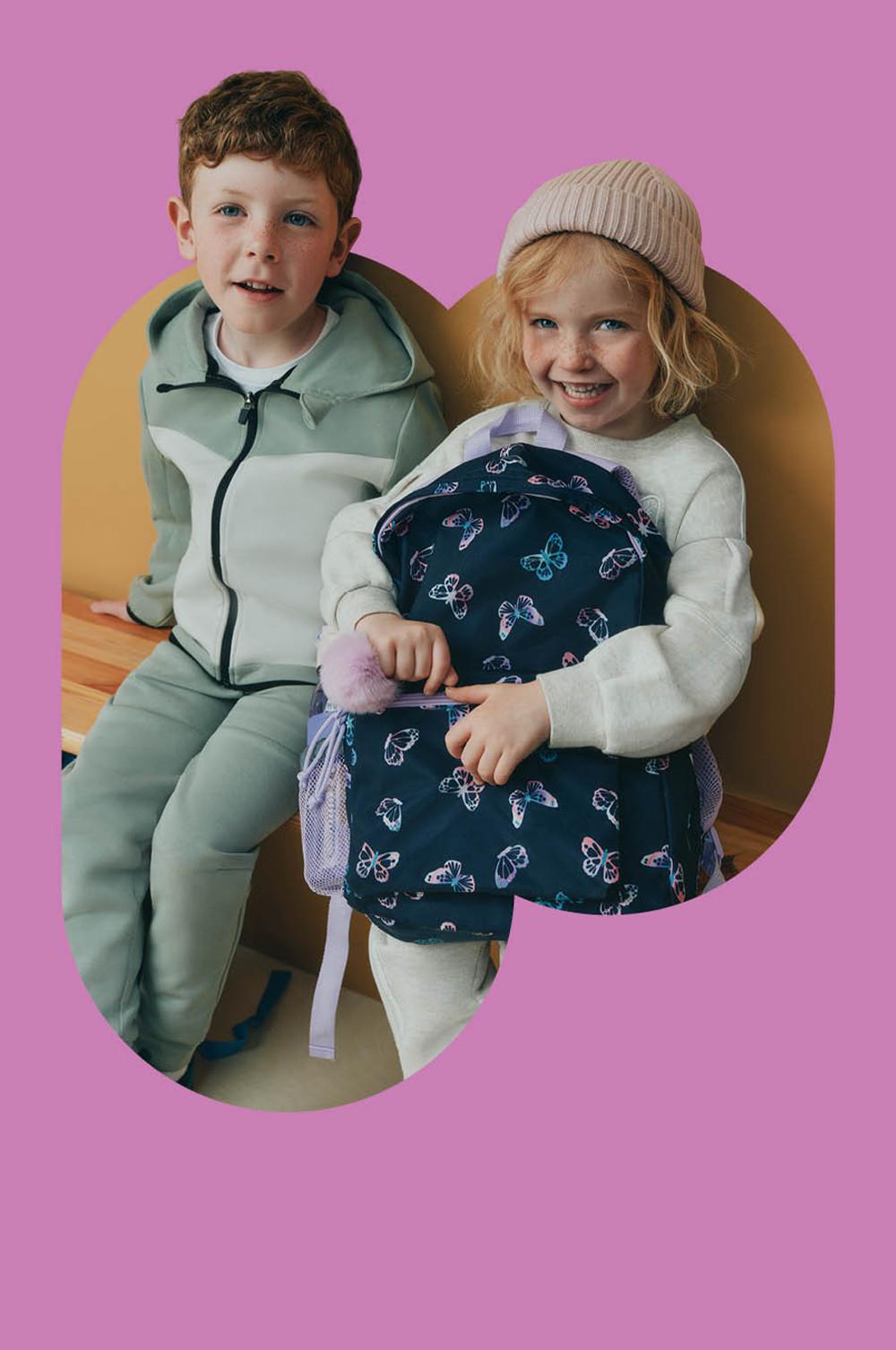 Two children wearing casual clothing. One in mint tracksuit, other in white sweater with knit beanie and butterfly print backpack.