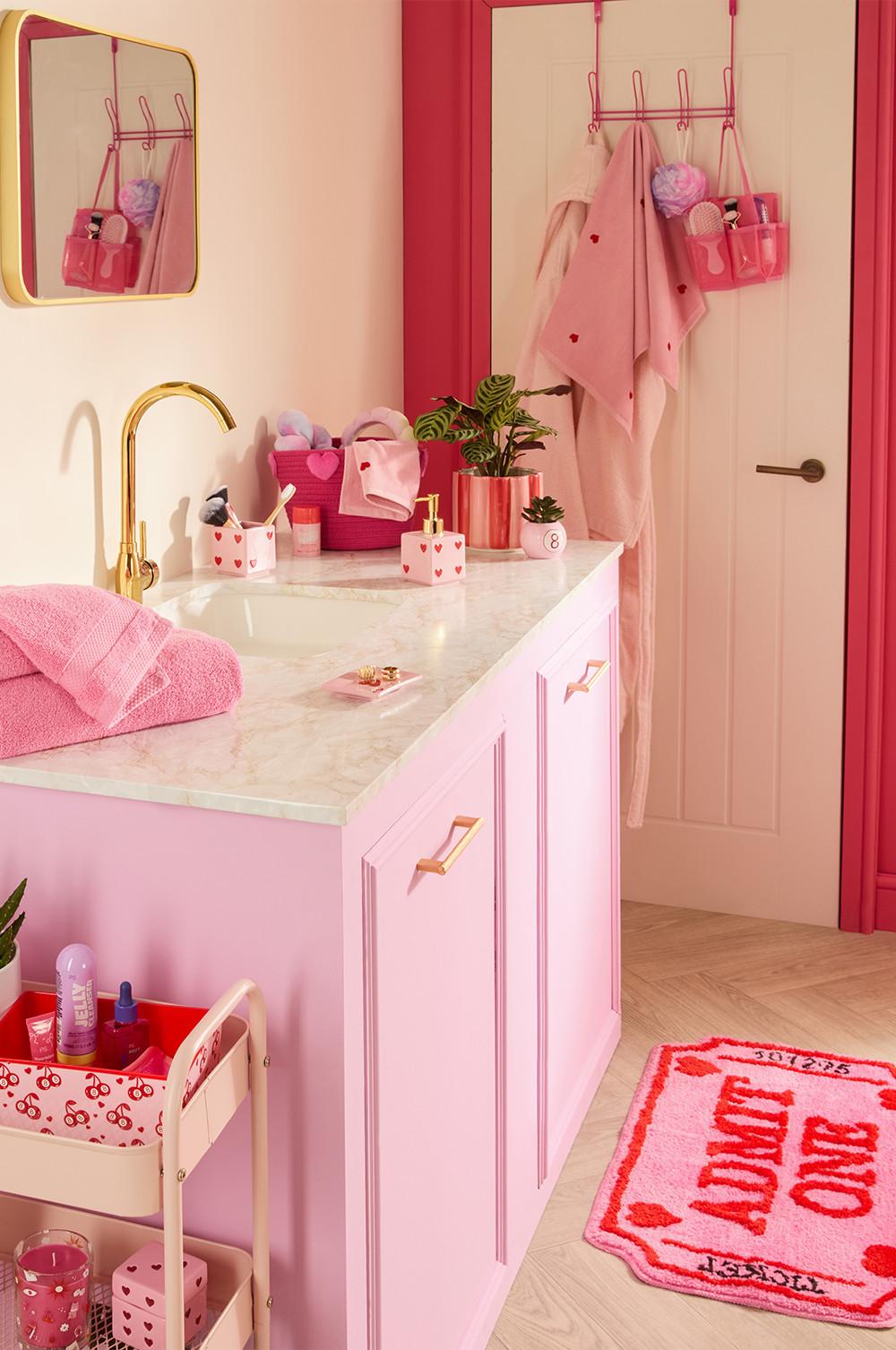 Salle de bain rose avec plan effet marbre, robinet doré et accessoires à cœurs. Des serviettes sont accrochées à des crochets de porte, avec un petit panier de bain visible.
