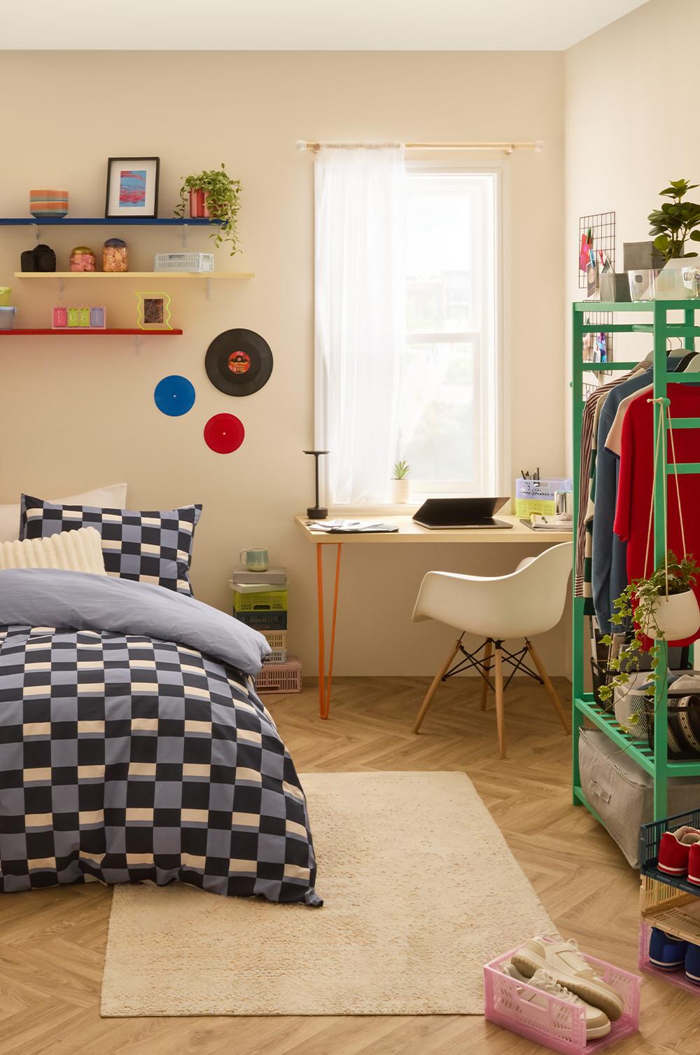 Chambre stylée avec linge de lit à carreaux, coin bureau, chaise blanche et étagère verte contenant vêtements et plantes.