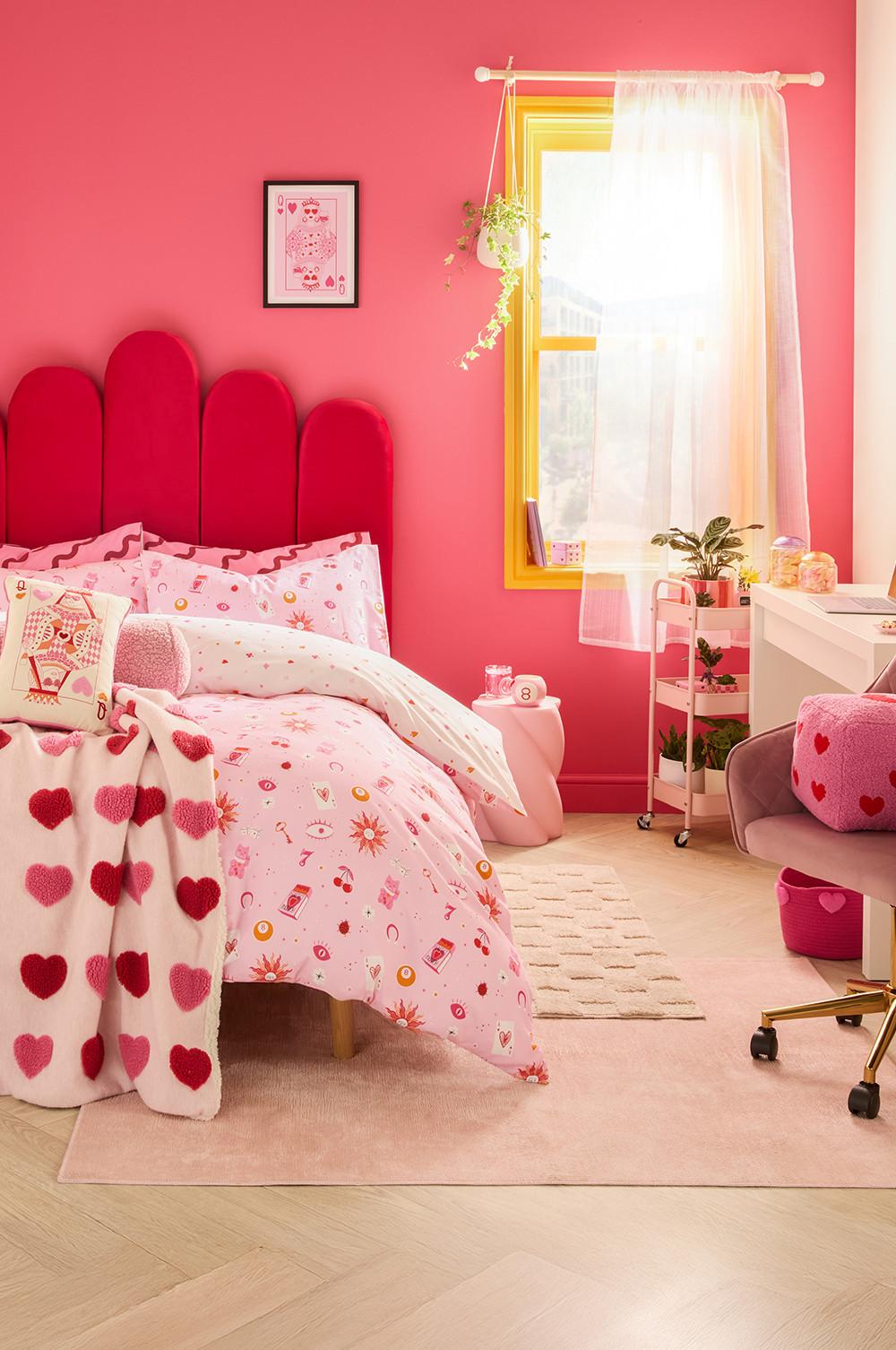 Chambre rose avec une tête de lit rouge festonnée, du linge de lit et un plaid à motifs de cœurs. Fenêtre avec châssis jaune, un rideau blanc et une plante suspendue. Murs roses et coin bureau.