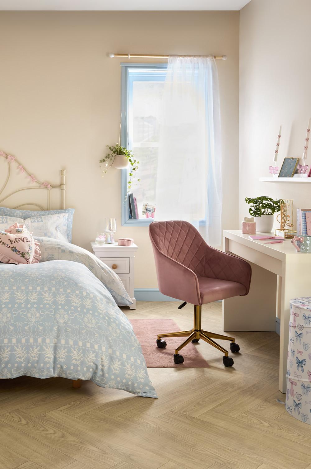 Chambre stylée avec chaise de bureau rose matelassée et base dorée, bureau blanc et parure de lit bleu clair complétée par des coussins décoratifs.
