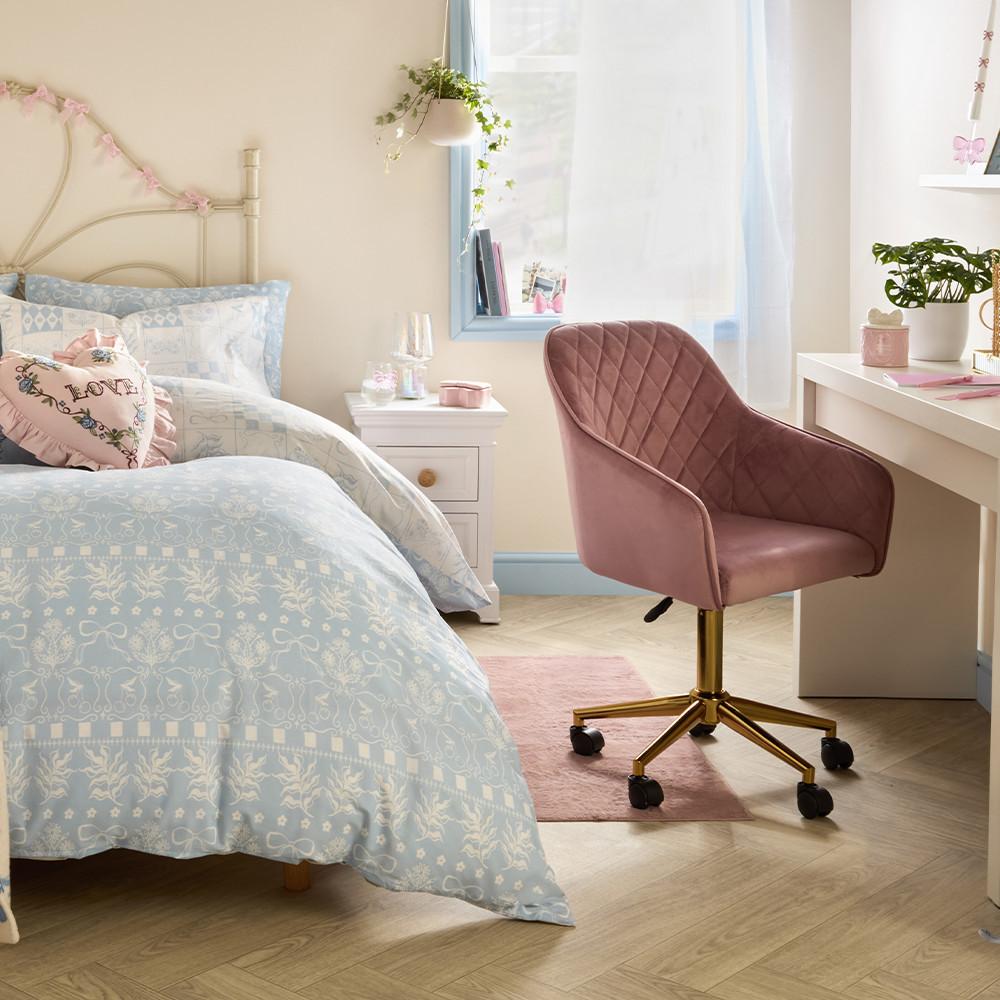 Linge de lit bleu clair à motifs avec coussin rose en forme de cœur, table de chevet blanche et chaise en velours rose dans une chambre.