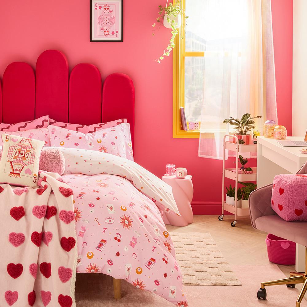Habitación rosa con cabecero rojo festoneado, ropa de cama con estampado de corazones y decoración a juego, incluyendo un espejo con marco amarillo y muebles rosa.
