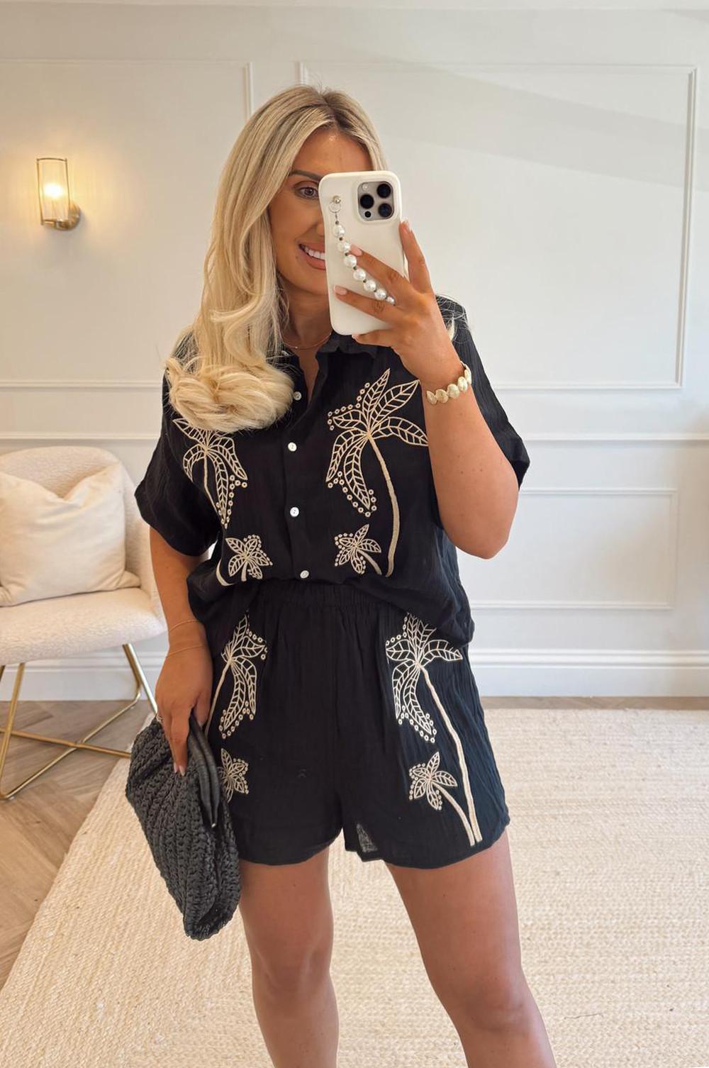 Model wearing black button-up shirt with embroidered palm tree motifs and matching shorts, paired with a dark woven handbag.