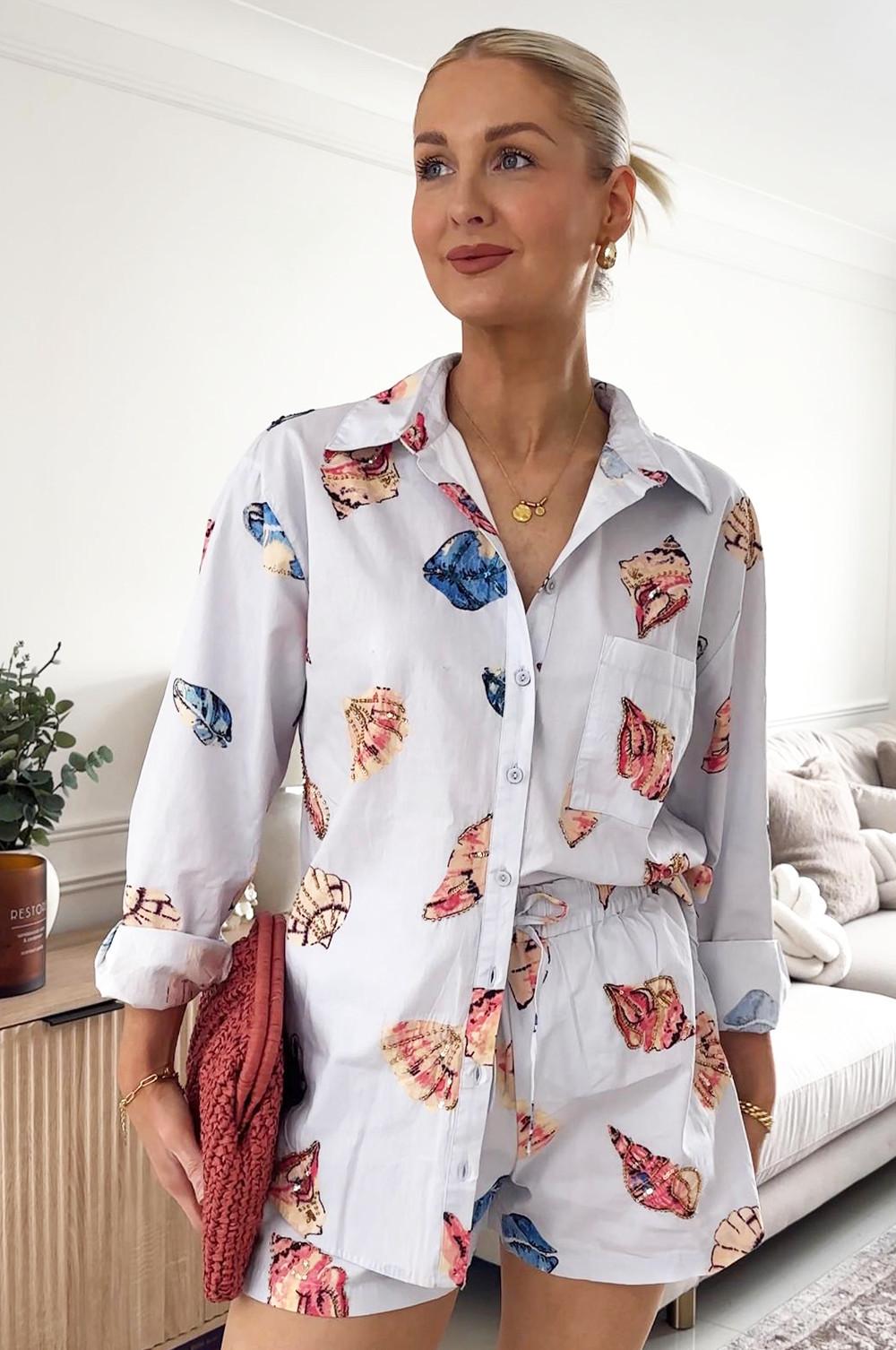 Model wearing light blue button-up shirt with colourful seashell pattern paired with matching shorts. Gold-toned necklace accessories and a red woven bag,