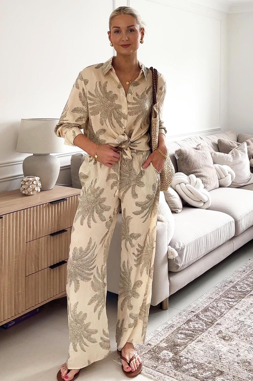 Model wearing a cream and beige tropical print co-ord set with knotted shirt and wide leg trousers, styled with a woven bag.