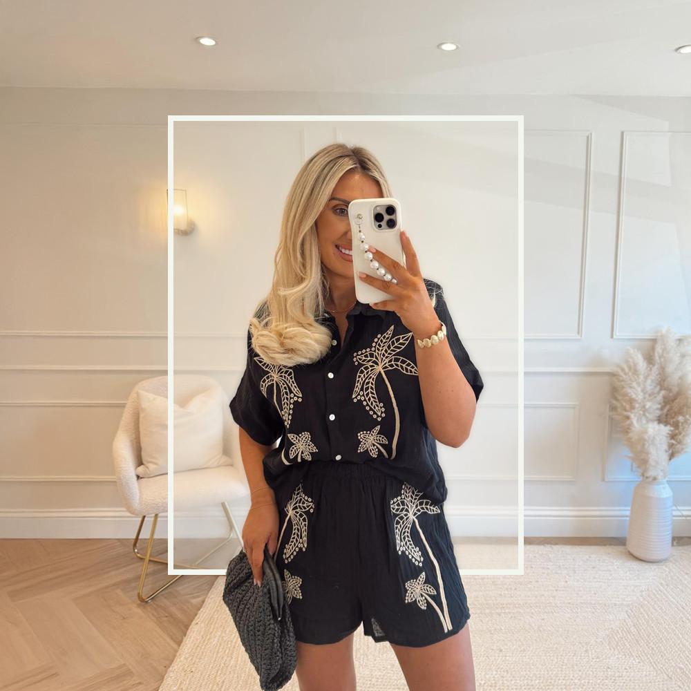 Modle wearing black button-up shirt and shorts co-rd with embroidered palm tree motifs, paired with a dark woven handbag.