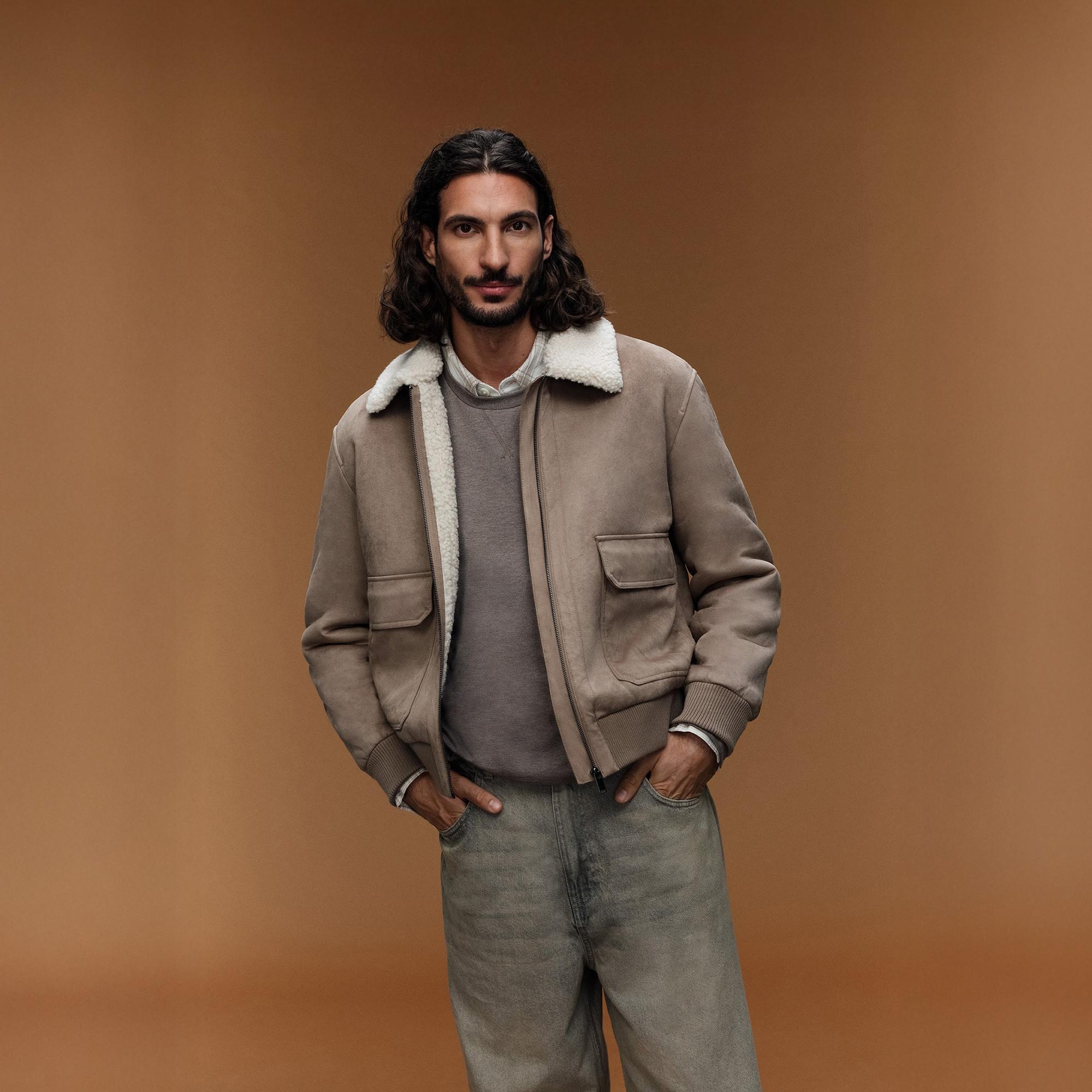 Bomber beige con collo in montone shearling abbinato a maglione grigio e jeans chiari su uno sfondo neutro.
