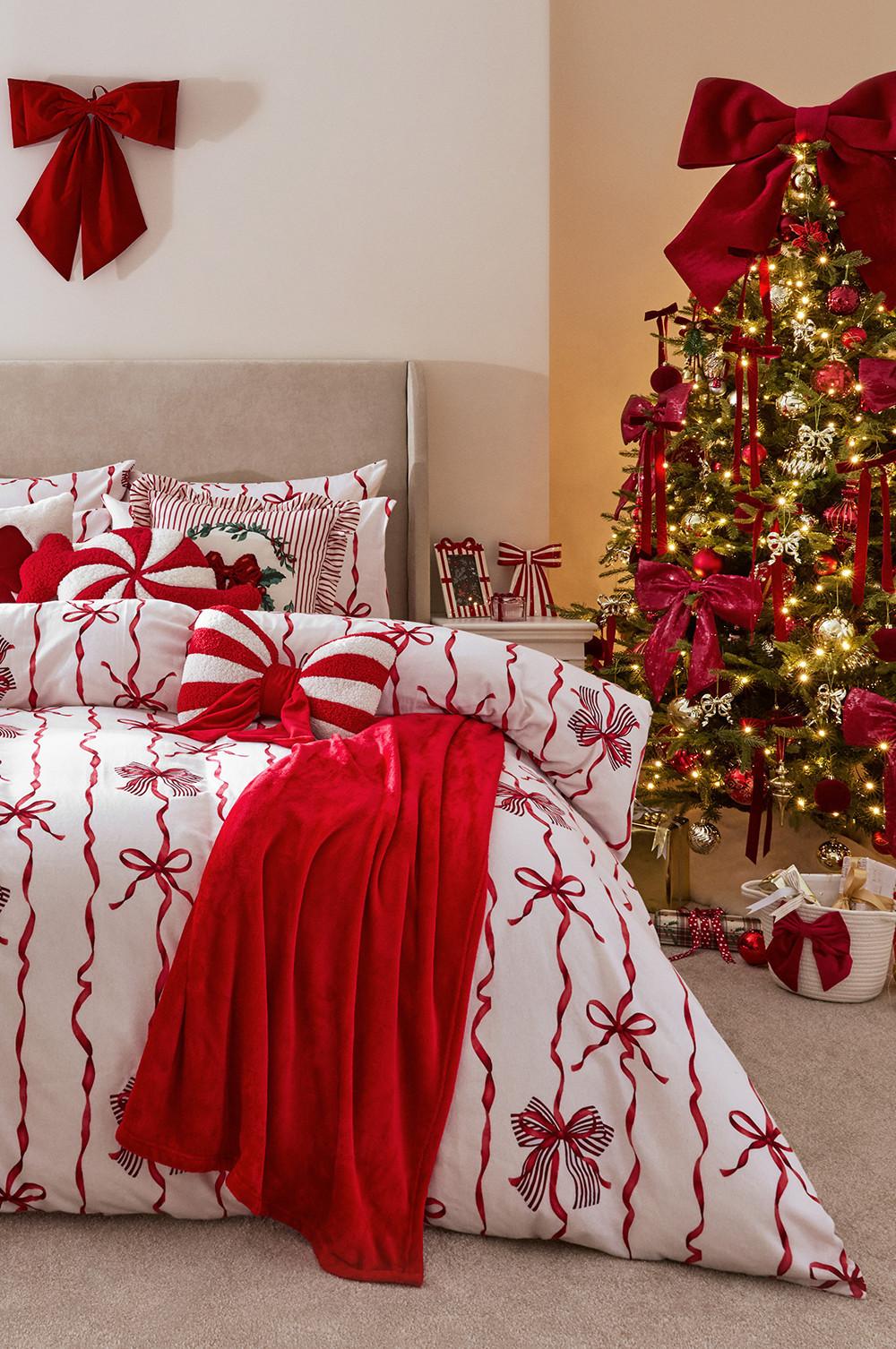 Camera da letto decorata per le feste con biancheria da letto a fantasia con fiocchi rossi e bianchi accanto a un albero di Natale decorato con nastri rossi.