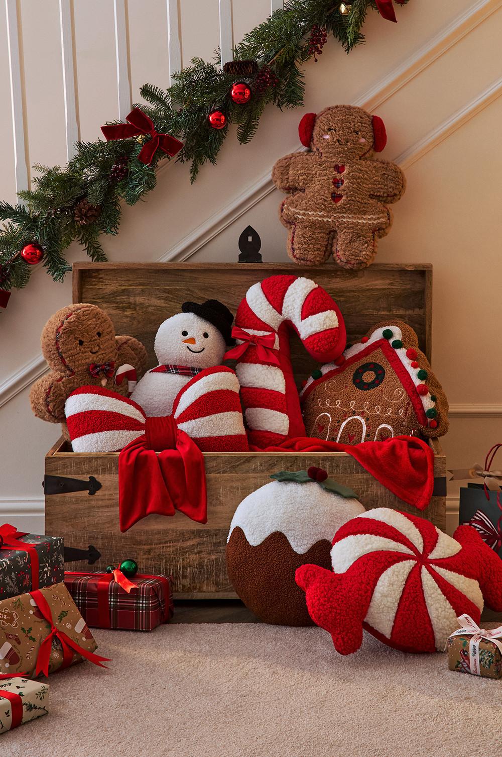 Decorazioni natalizie festive tra cui peluche, cuscini a forma di bastoncino di zucchero e di pan di zenzero in un baule di legno con ghirlanda al di sopra.
