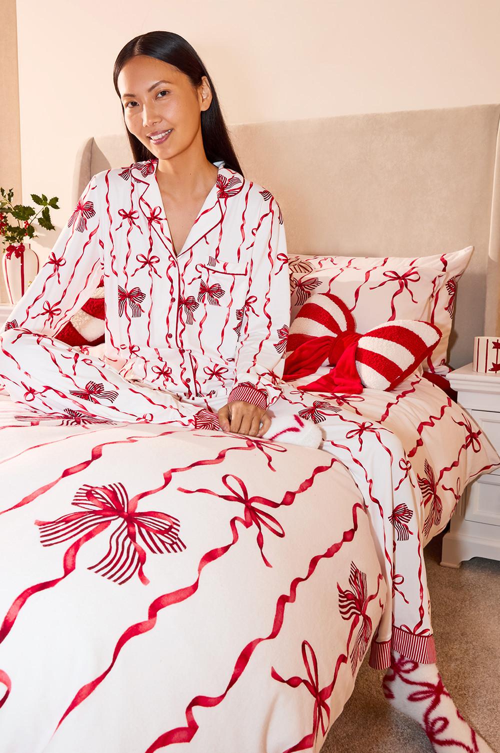 Persona che indossa un pigiama festivo bianco e rosso con motivo a fiocchi, seduta su un letto coordinato con cuscini decorativi.