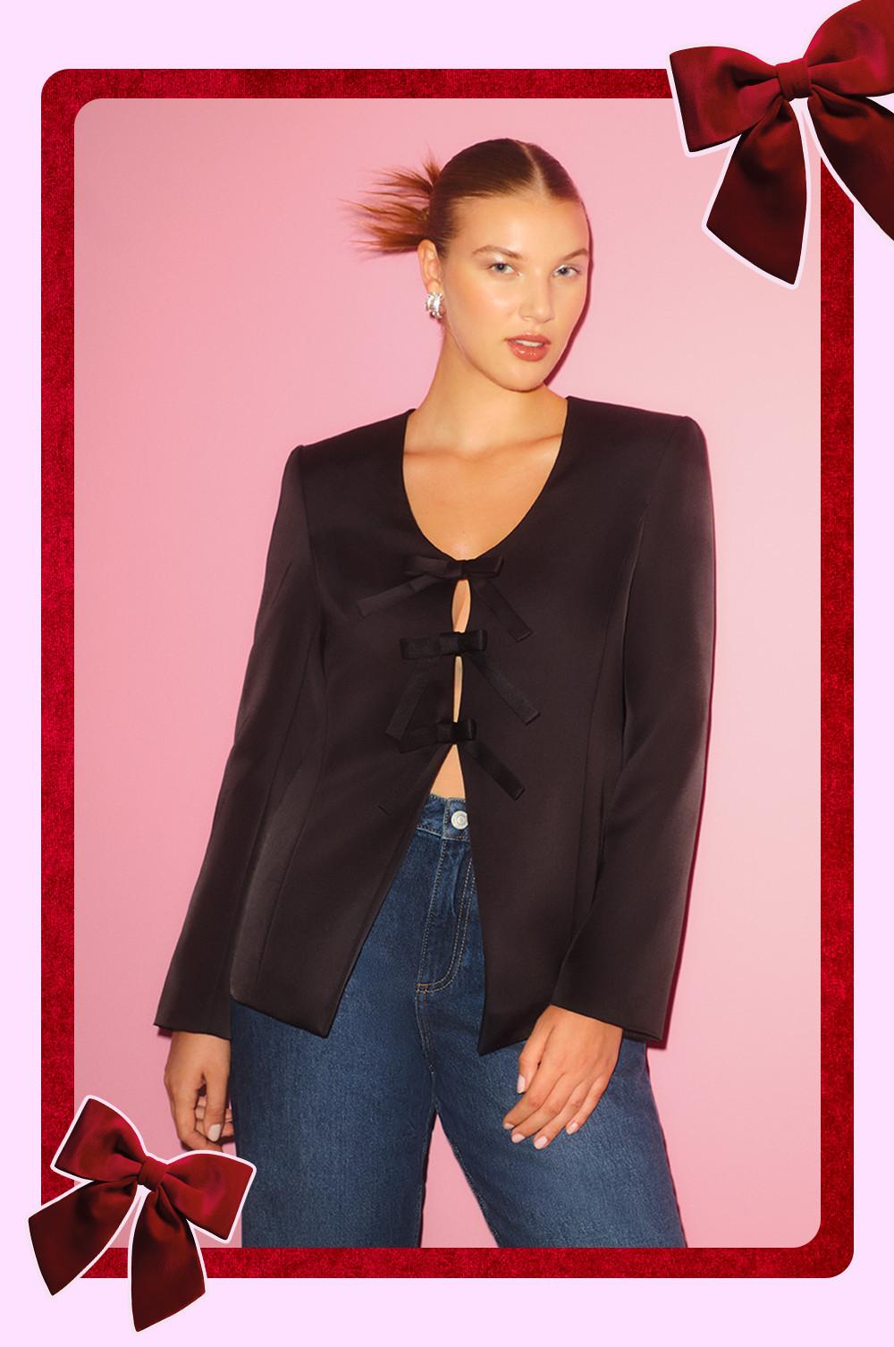 Giacca nera con chiusura a fiocco indossata con jeans blu, incorniciata da un bordo con nastro rosso su sfondo rosa.