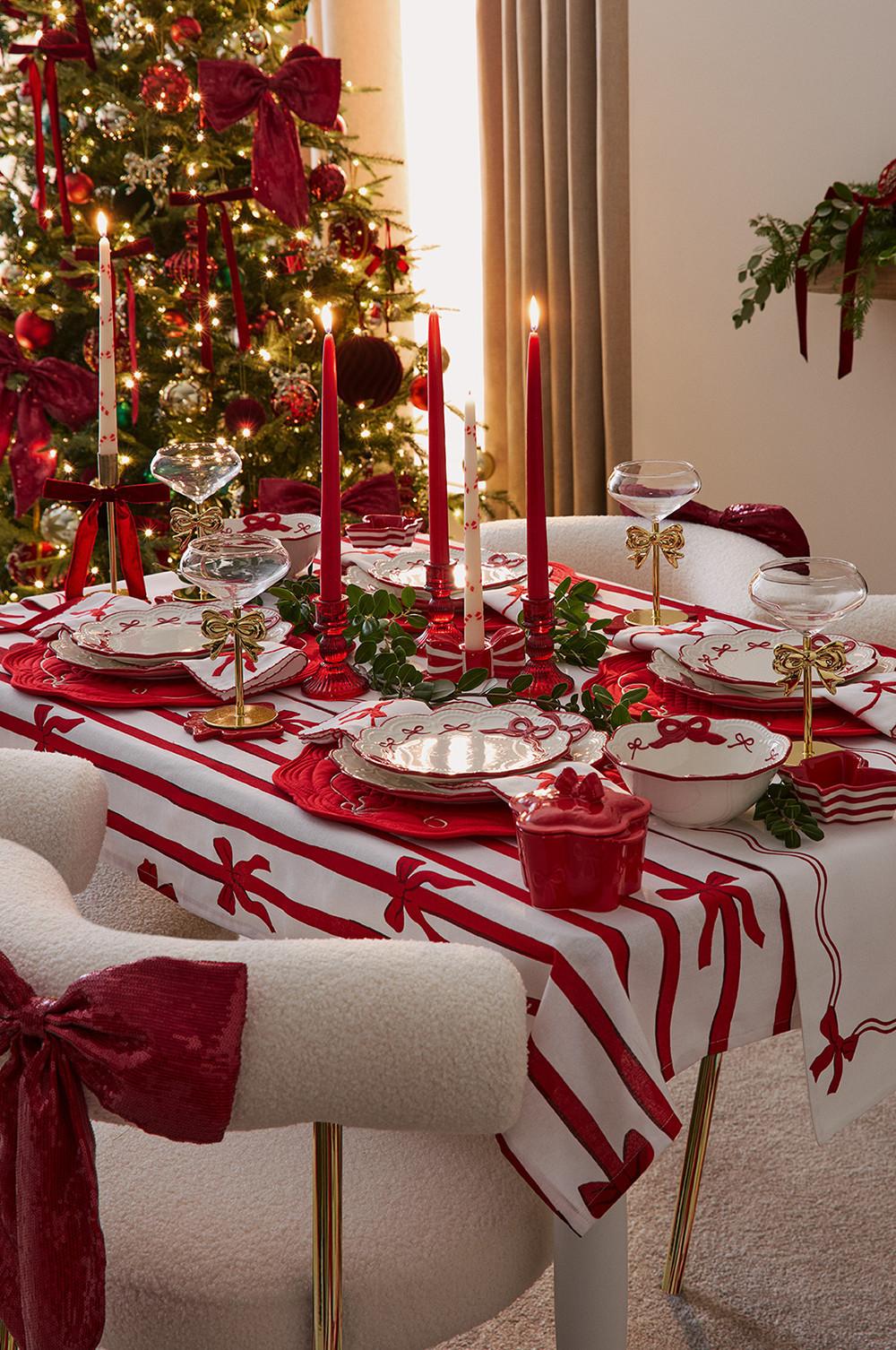 Tavola imbandita a festa con stoviglie rosse e bianche, candele accese e bicchieri per lo champagne accanto a un albero di Natale addobbato.