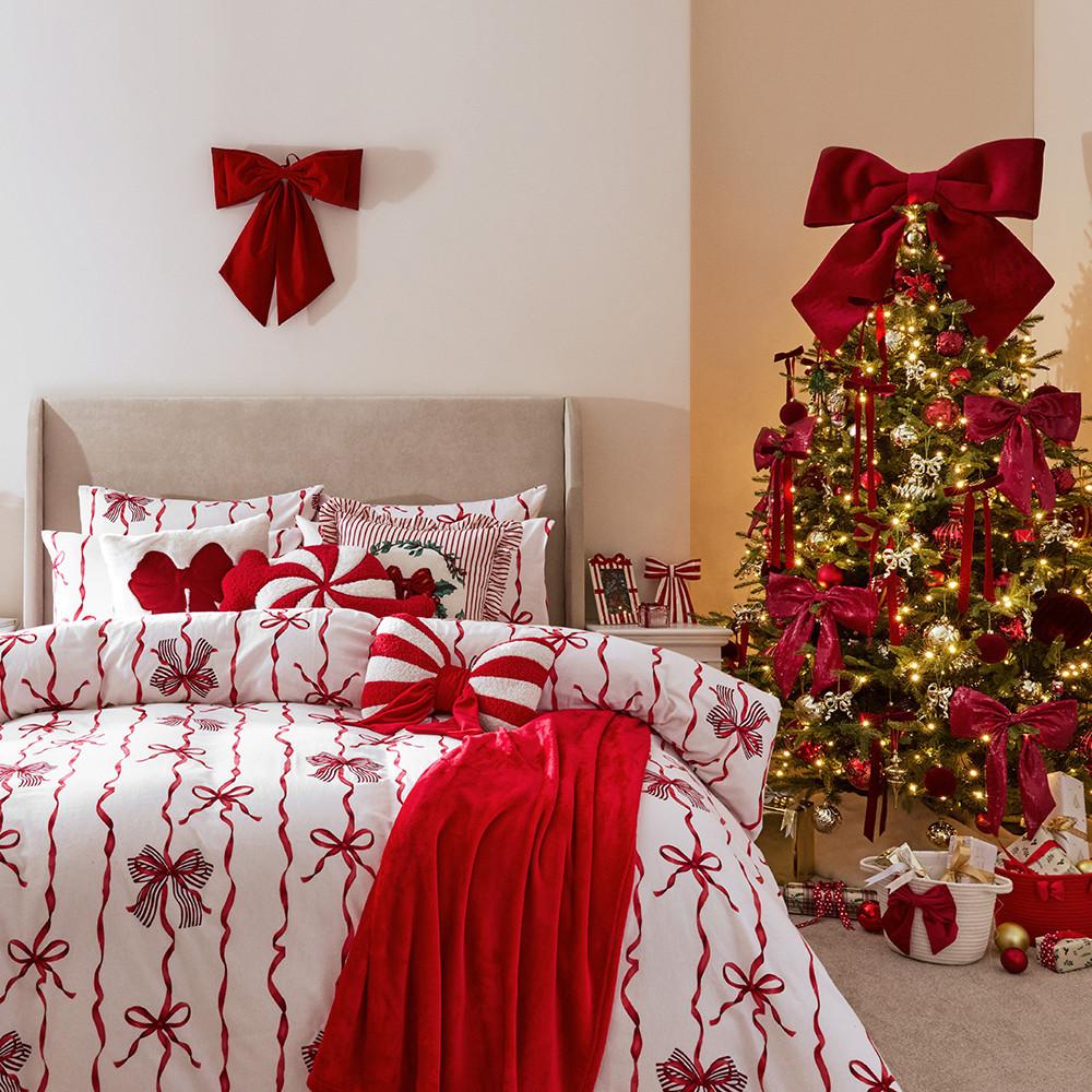 Camera da letto decorata per le feste con biancheria da letto a fantasia con fiocchi rossi e bianchi, cuscini decorativi e albero di Natale con nastri rossi e luci.