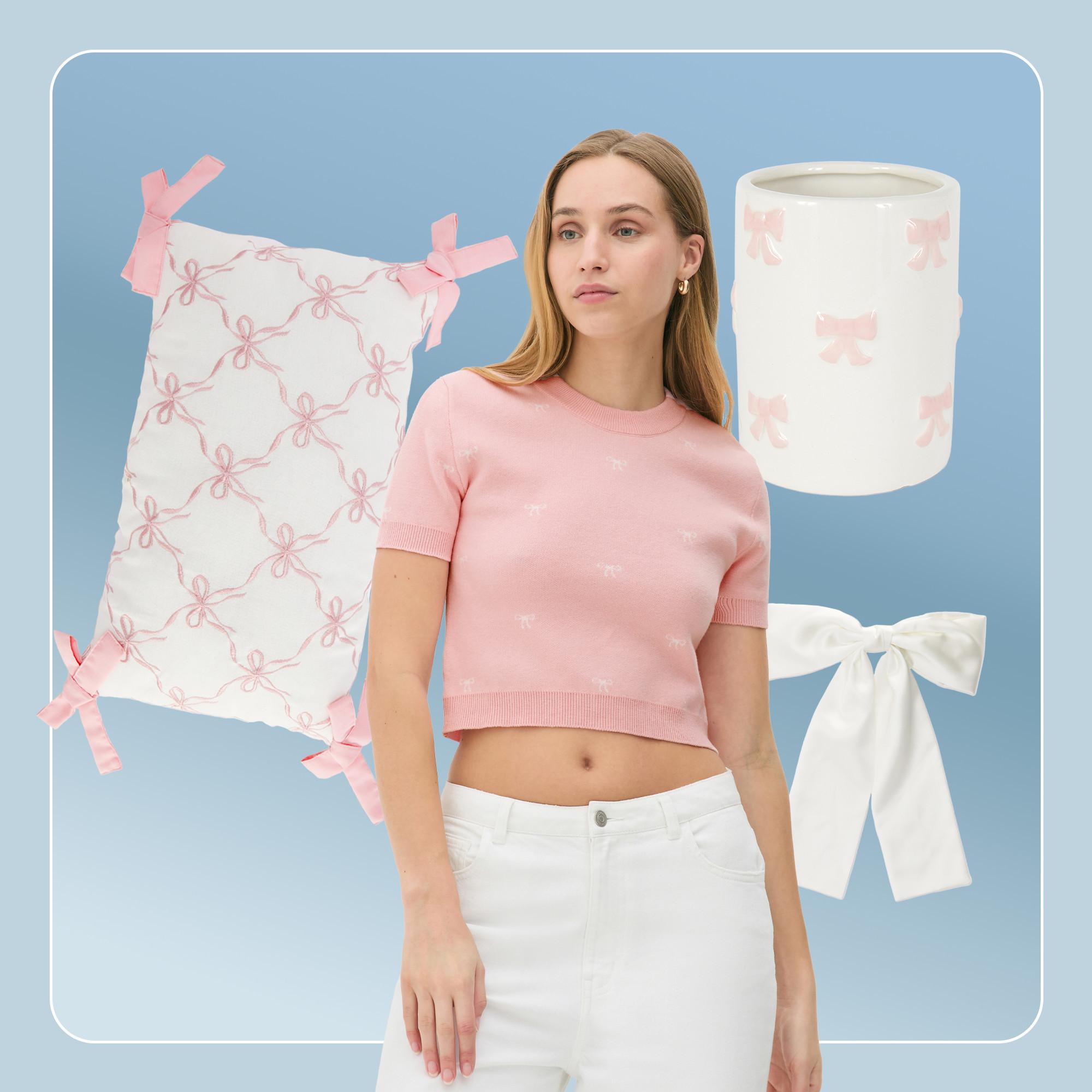 Model wearing pink cropped knit top and white jeans. White pillow and bathroom tumbler with pink bow print in background.