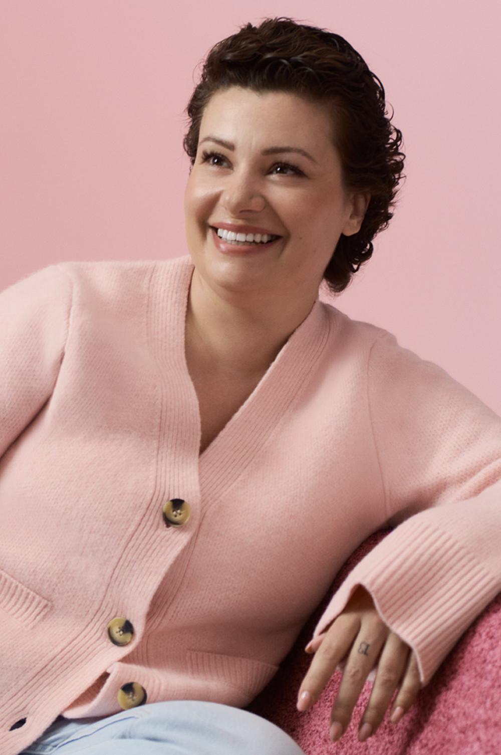 Persona con capelli ricci che indossa un cardigan rosa chiaro con bottoni in legno su uno sfondo rosa.