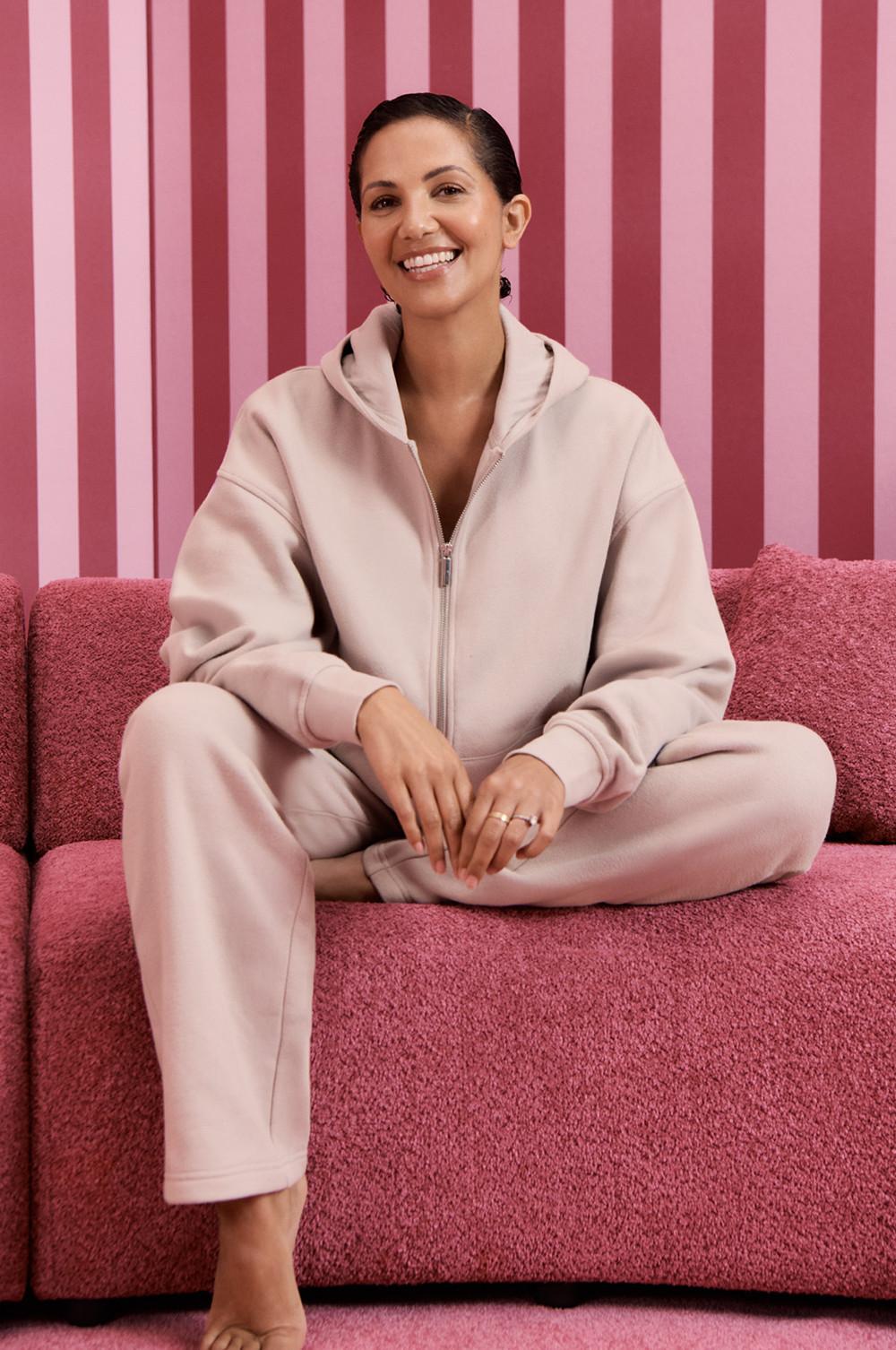 Persona che indossa un completo di abbigliamento da casa rosa chiaro con zip, seduta su un divano rosa su uno sfondo a righe.