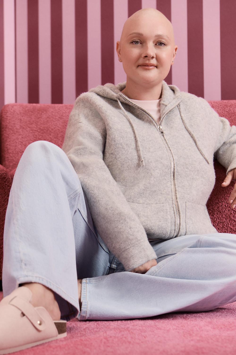 Persona che indossa una felpa grigia con cappuccio e zip e jeans denim chiari, seduta su un divano rosa su uno sfondo a righe.