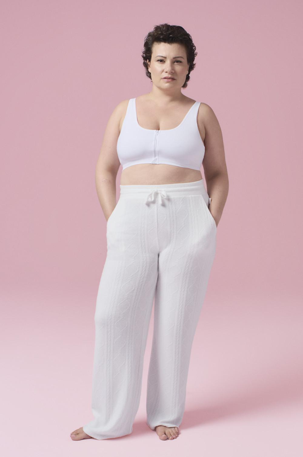 Persona che indossa un reggiseno sportivo bianco con zip frontale abbinato a pantaloni da casa bianchi con coulisse su uno sfondo rosa.