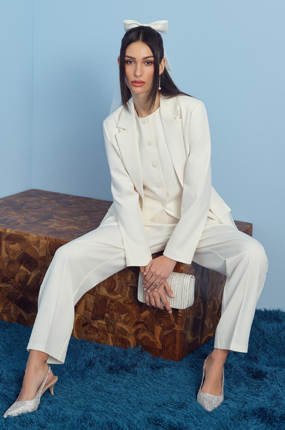 Model wearing white vest, blazer, pants, bow and rhinestone heels with clutch bag.