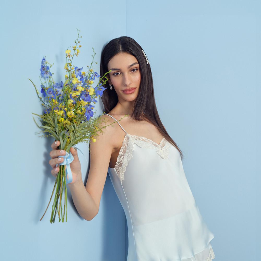 Model wearing a white satin camisole with lace detail, holding flowers.