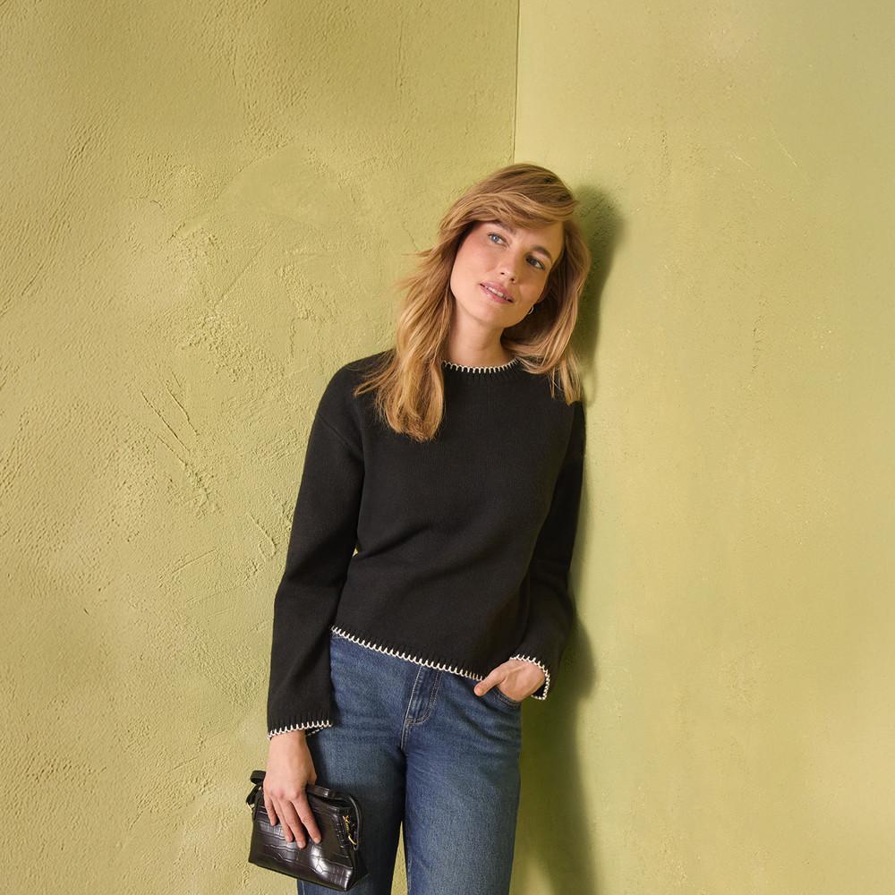 Black sweater with white contrast stitching along edges, paired with blue jeans and holding a small handbag.