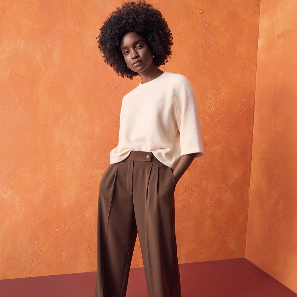 Person wearing cream short-sleeved top and brown wide leg trousers against an orange backdrop.