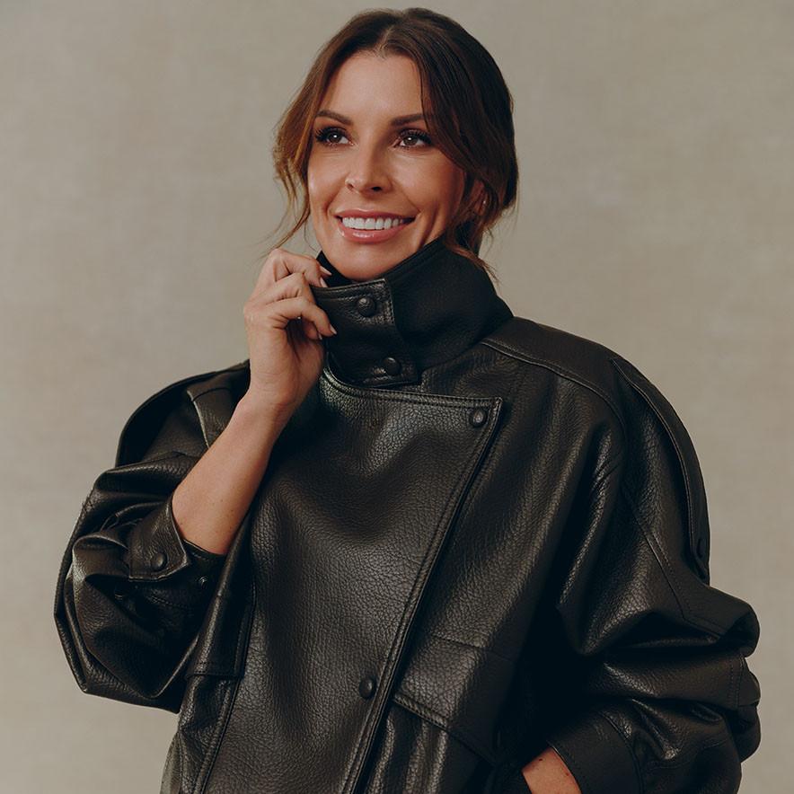 A woman with brown hair wears a dark brown, textured leather jacket with a high collar.