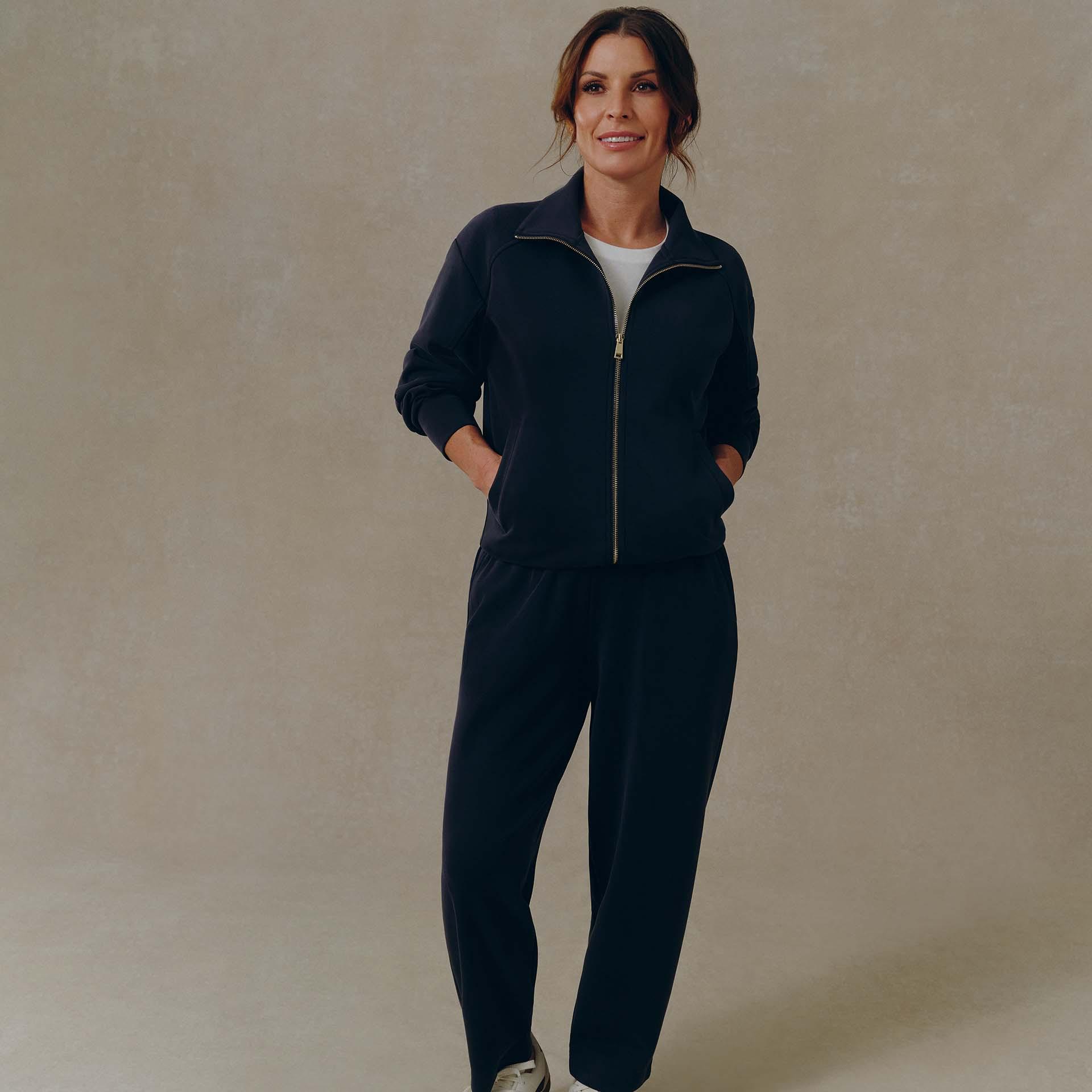 A woman with brown hair is wearing a dark navy zip-up tracksuit, with her hands in her pockets.
