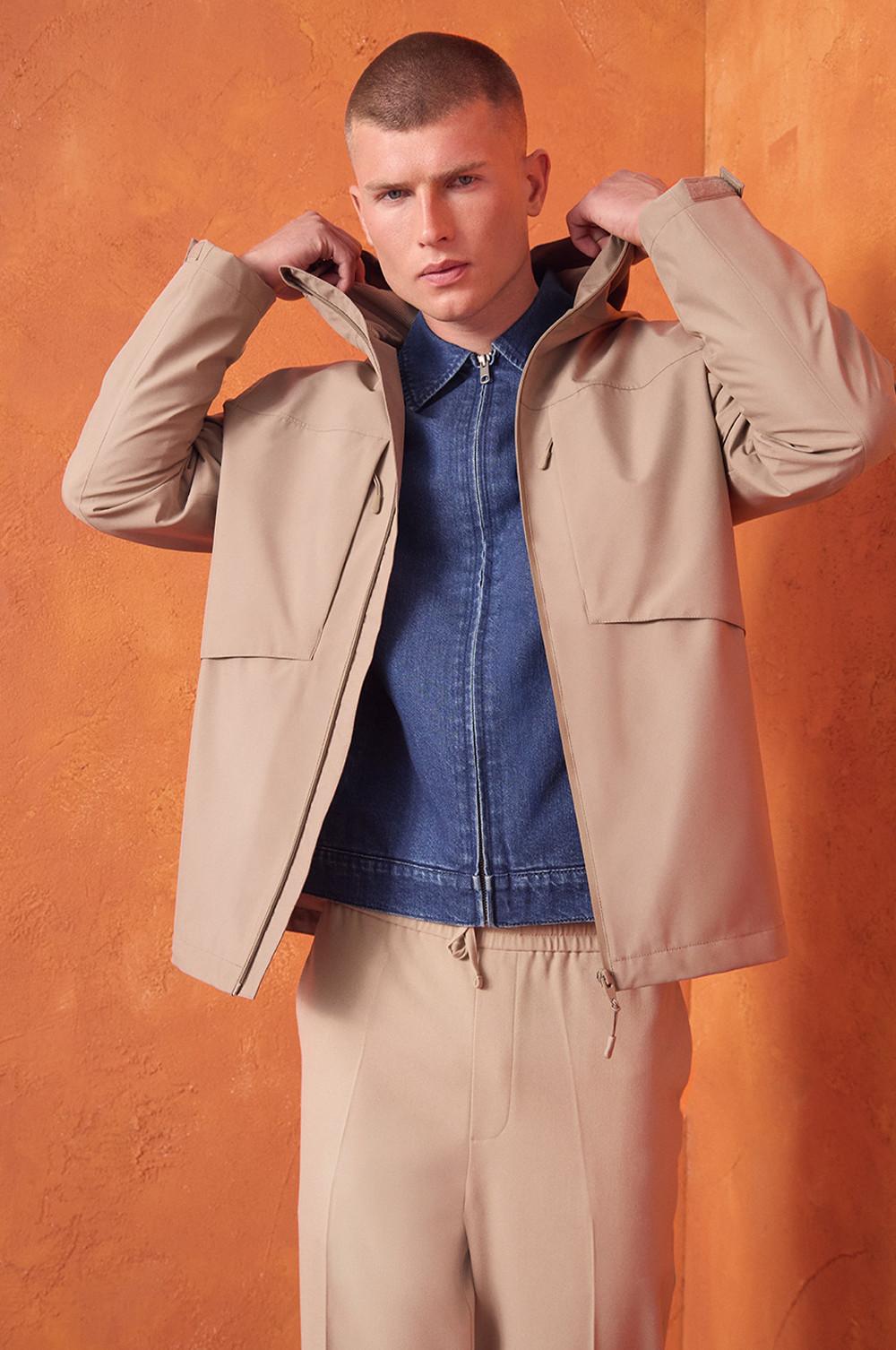 Persona che indossa una giacca beige su camicia di jeans e pantaloni beige abbinati davanti a un muro arancione.
