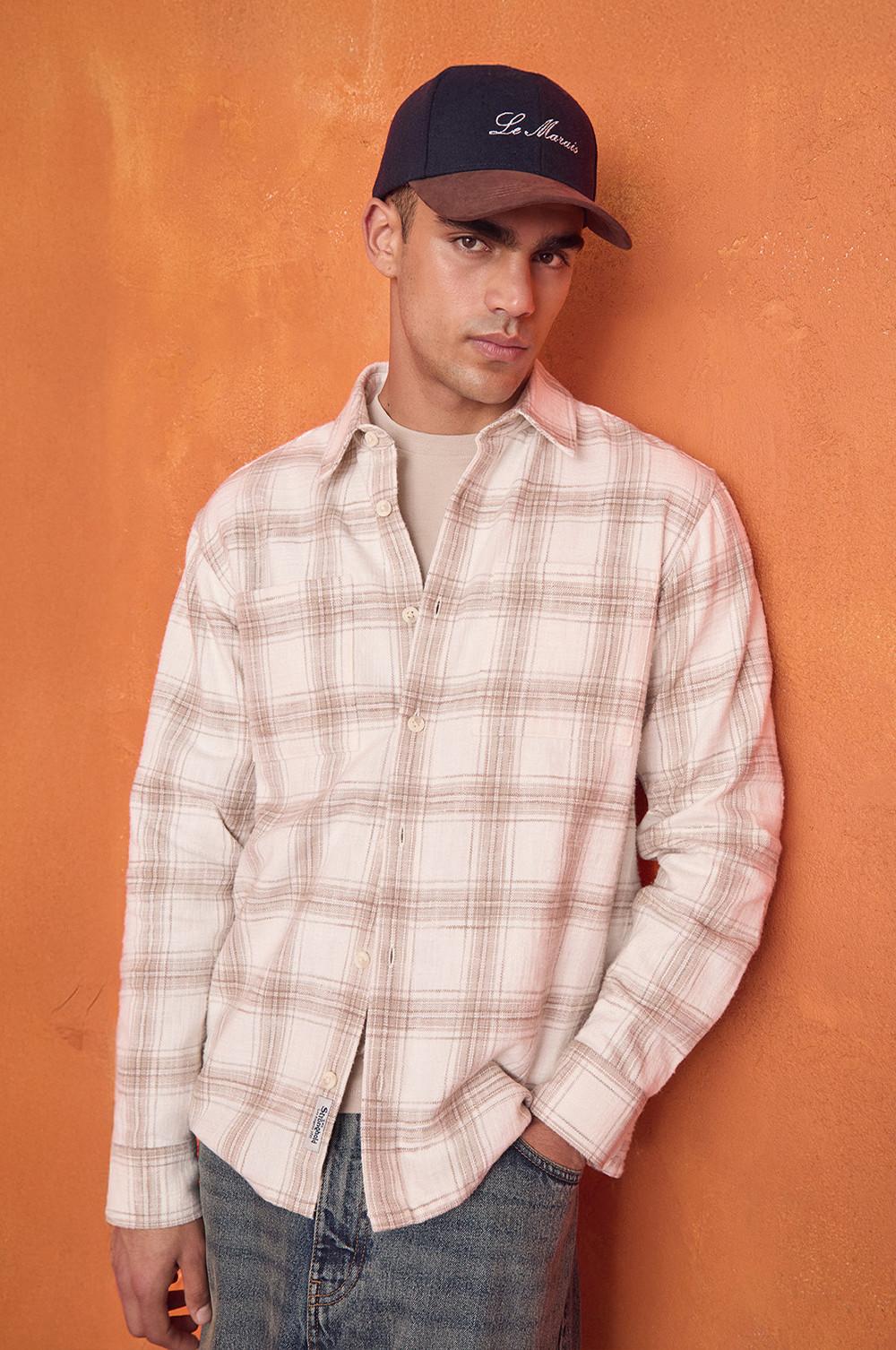 Cappellino "Le Marais" marrone e blu, indossato con una camicia a quadri rosa e panna su una maglietta bianca e dei jeans su uno sfondo arancione.
