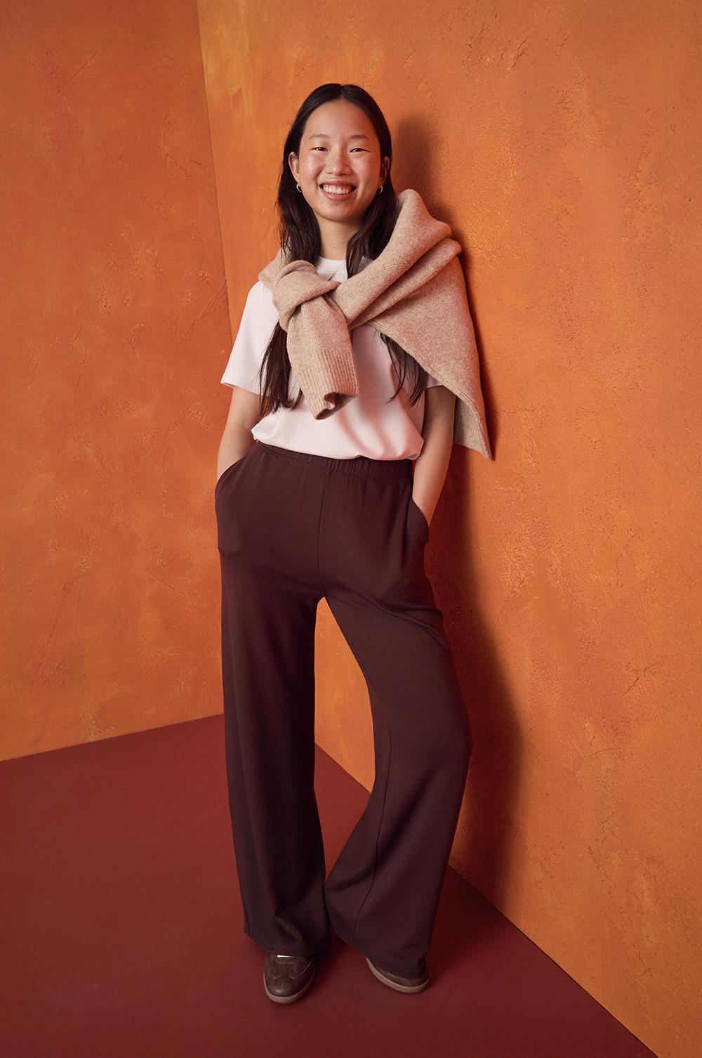 Persona che indossa una maglietta bianca con pantaloni a gamba larga marroni e un maglione beige drappeggiato sulle spalle contro un muro arancione.