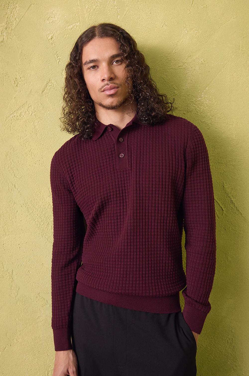 Maglia dolcevita a coste bordeaux con maniche lunghe e colletto, abbinata a pantaloni neri su una parete gialla ruvida.