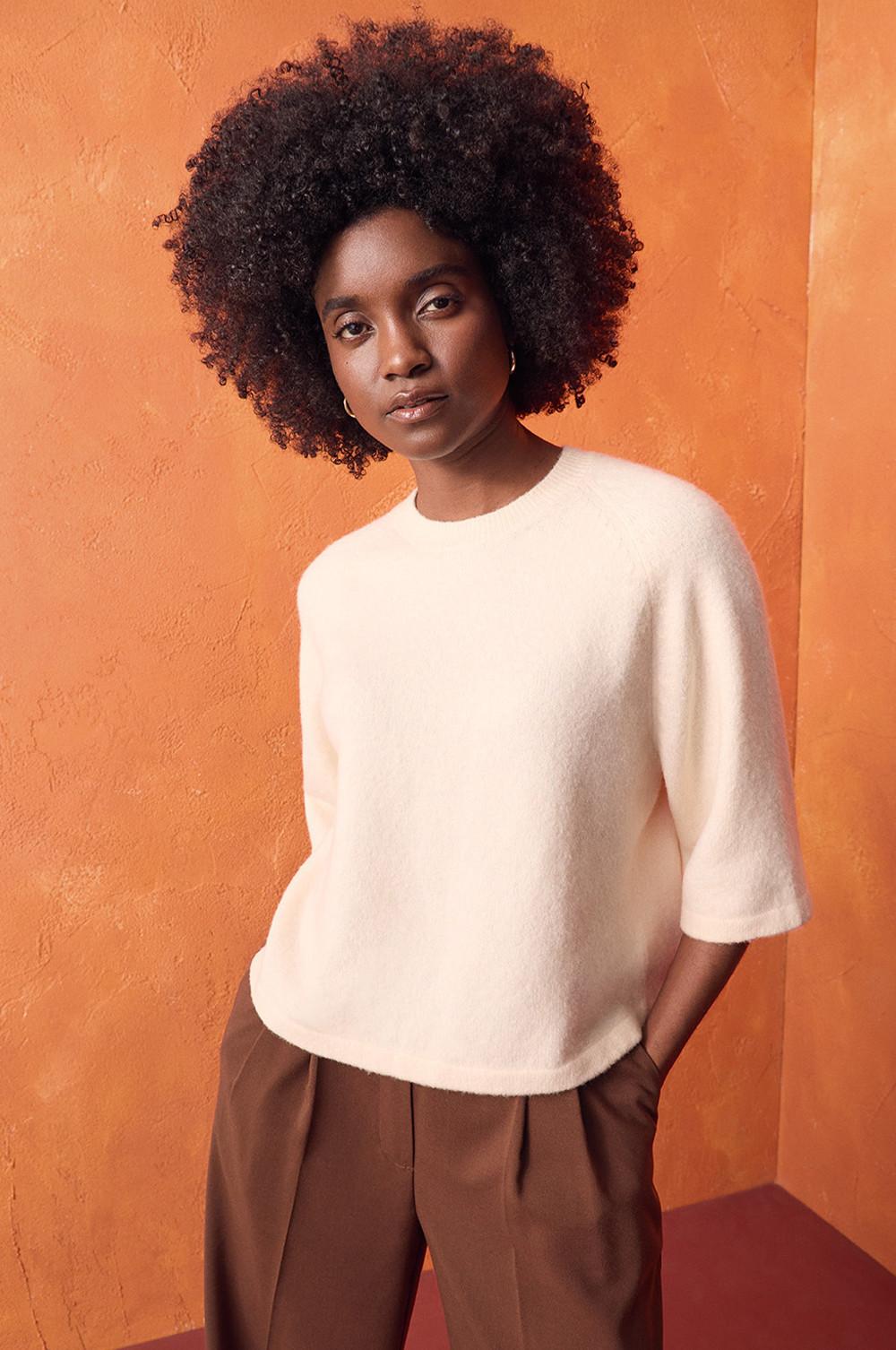 Persona che indossa un maglione a maniche corte color panna con pantaloni marroni su uno sfondo arancione.