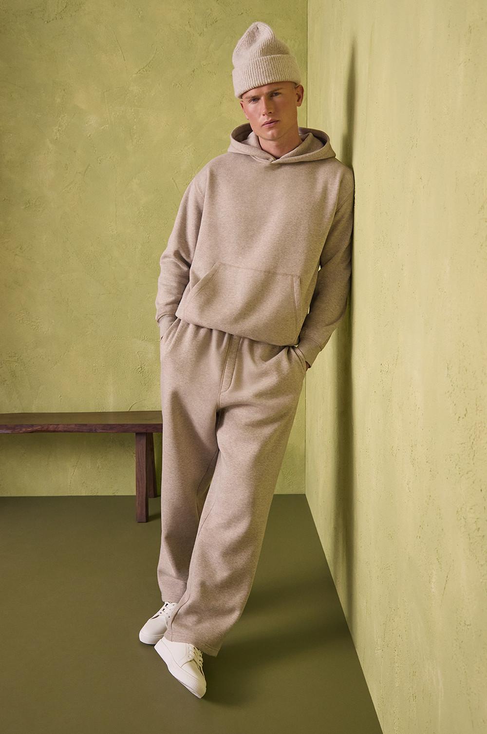 Set di abbigliamento da casa beige composto da felpa con cappuccio e tasca frontale abbinata a pantaloni della tuta e scarpe da ginnastica bianche, completato da un berretto in maglia.