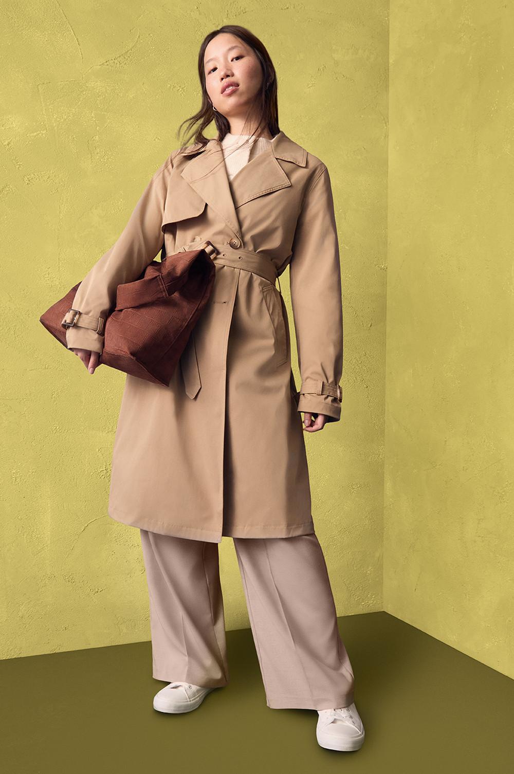 Trench beige con cintura indossato con pantaloni a gamba larga abbinati e scarpe da ginnastica bianche, completato da una borsa tote marrone.