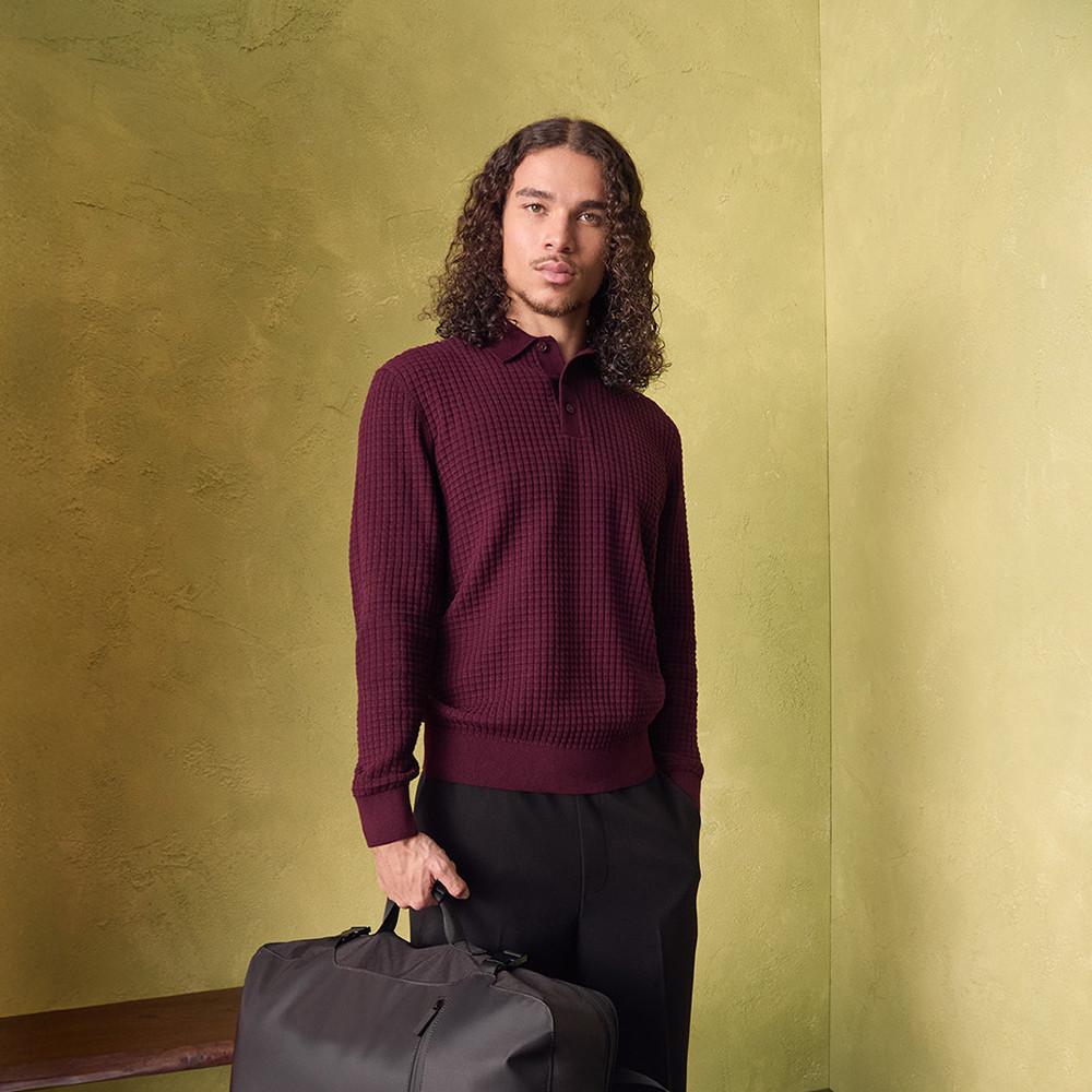 Persona che indossa una polo bordeaux con pantaloni neri e tiene in mano una borsa da viaggio grigia.