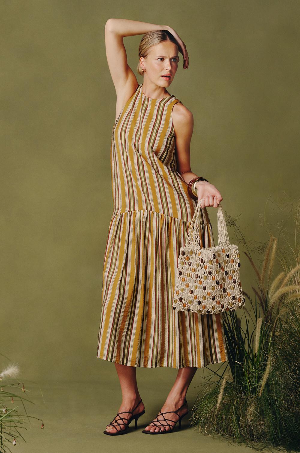 Femme tenant un sac crème en crochet et portant une robe mi-longue sans manches à rayures et jupe à volants avec des sandales à brides.