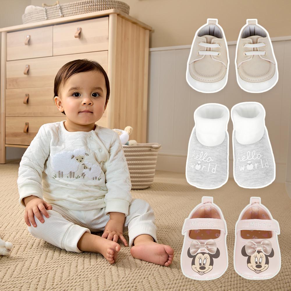 Baby in weißem Outfit sitzt auf einem Teppich mit drei Paar Babyschuhen: beige Schnürschuhe, graue „Hello World“-Stiefelchen und rosa Minnie-Maus-Schuhe.