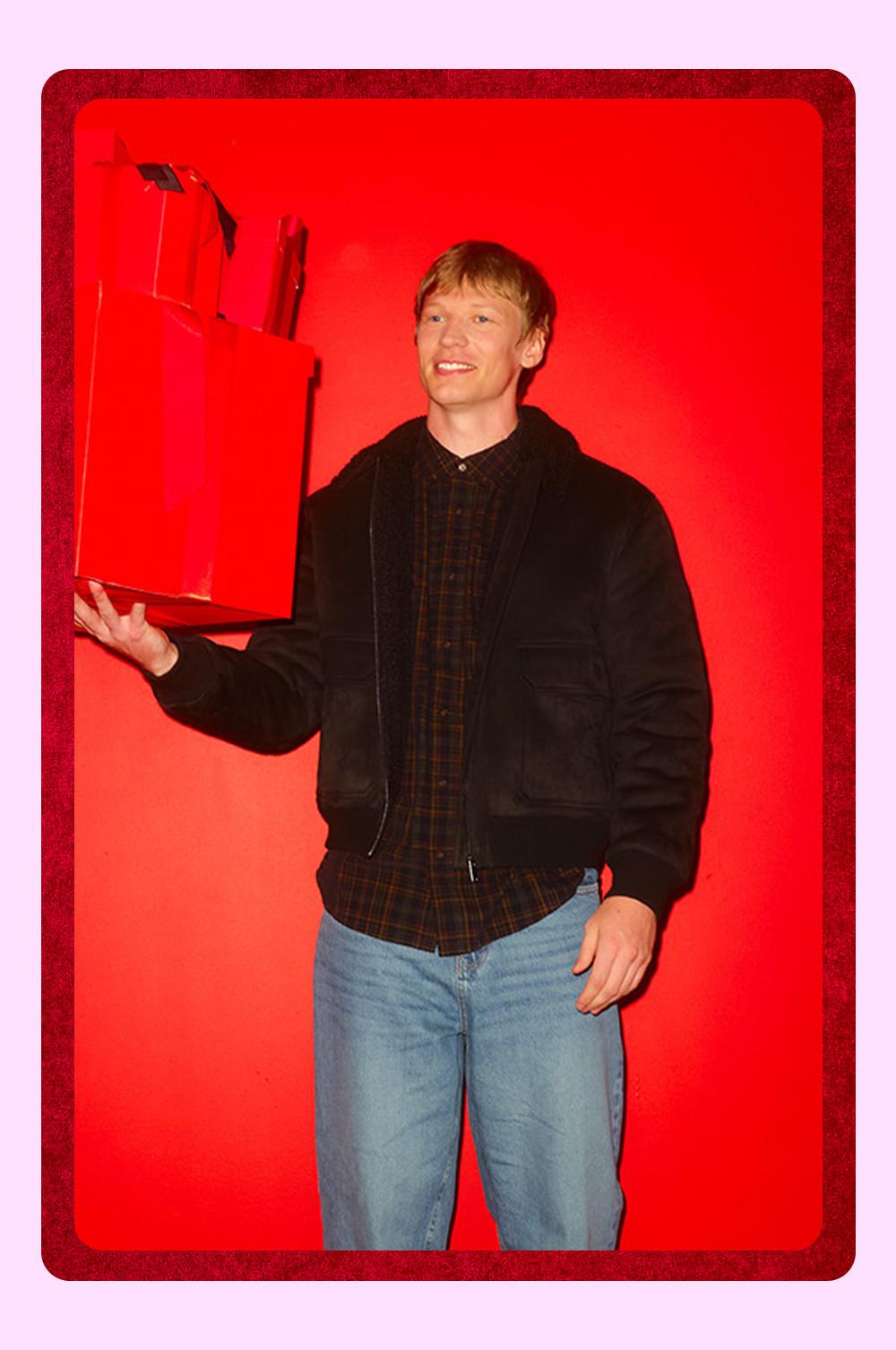 Persona che indossa una giacca nera sopra una camicia a quadri e tiene in mano una pila di scatole regalo rosso brillante su sfondo rosso.