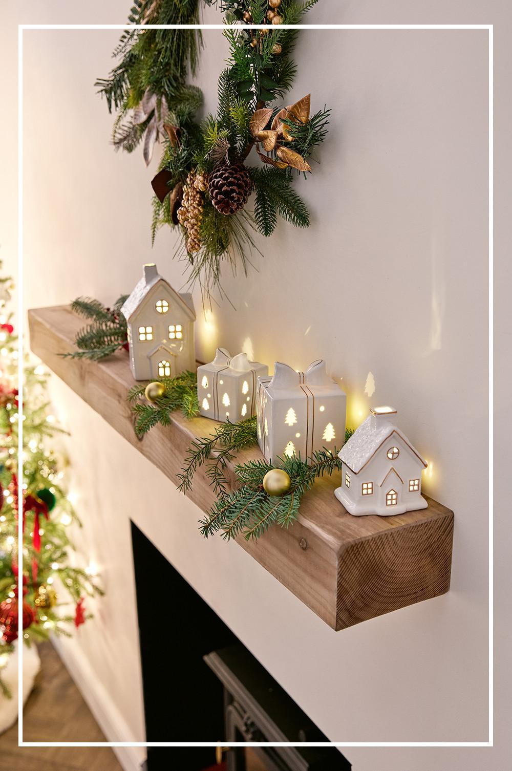 Wooden mantelpiece decorated with illuminated ceramic houses, gift boxes, pine branches, pinecones and baubles for Christmas.