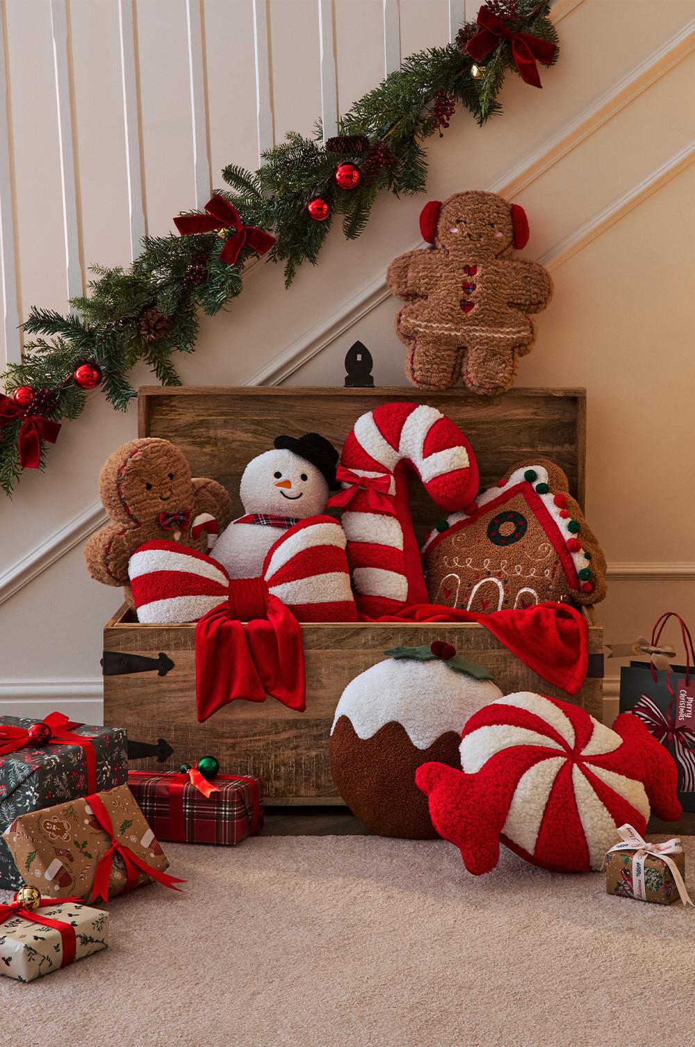 Wooden chest filled with festive plush toys including gingerbread men, snowman and candy cushions with wrapped presents nearby.