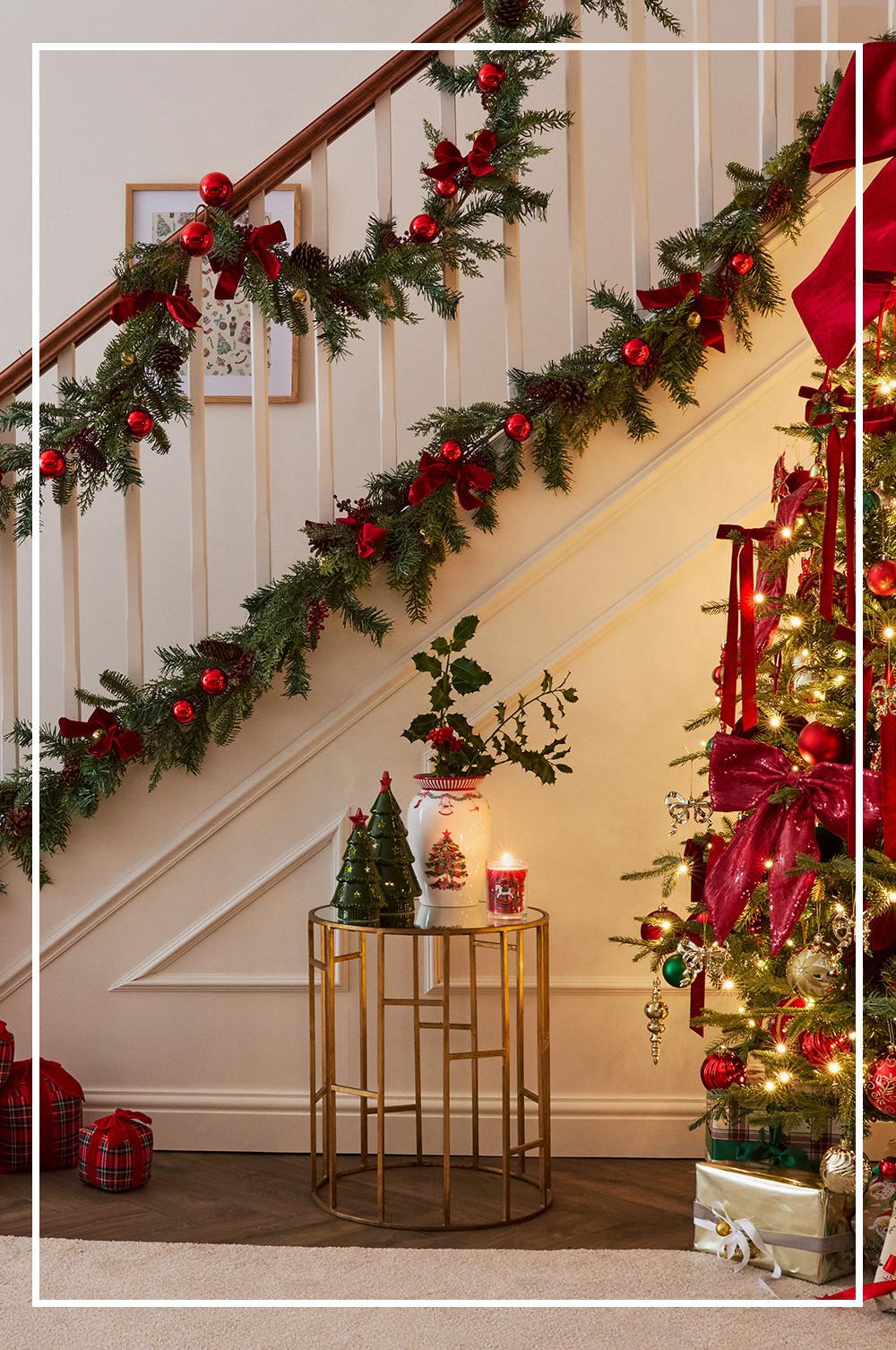Festive staircase decorated with evergreen garland, red baubles and ribbons, with Christmas tree and gold side table below.
