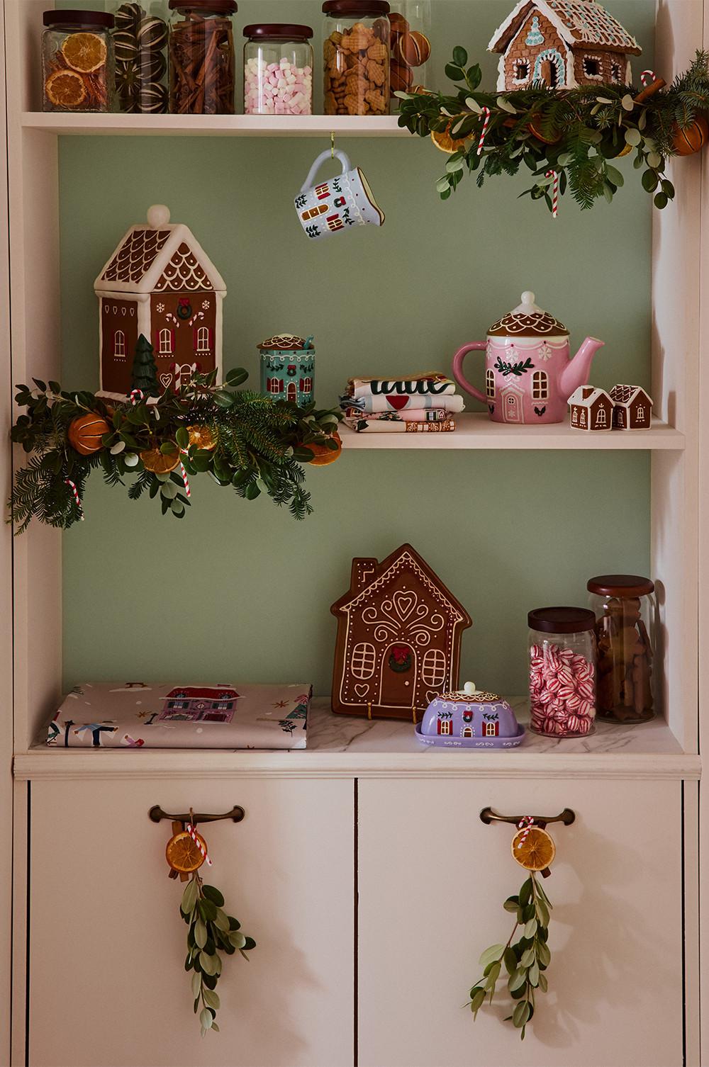 Festive shelving display with gingerbread houses, pink teapot, seasonal jars of sweets and dried oranges with evergreen garlands.