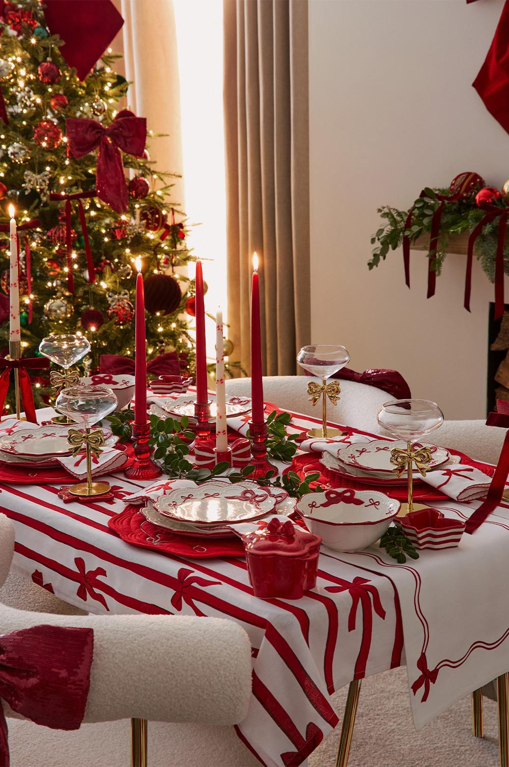 Festive dining table with red and white tablecloth, candles and glassware, beside decorated Christmas tree.