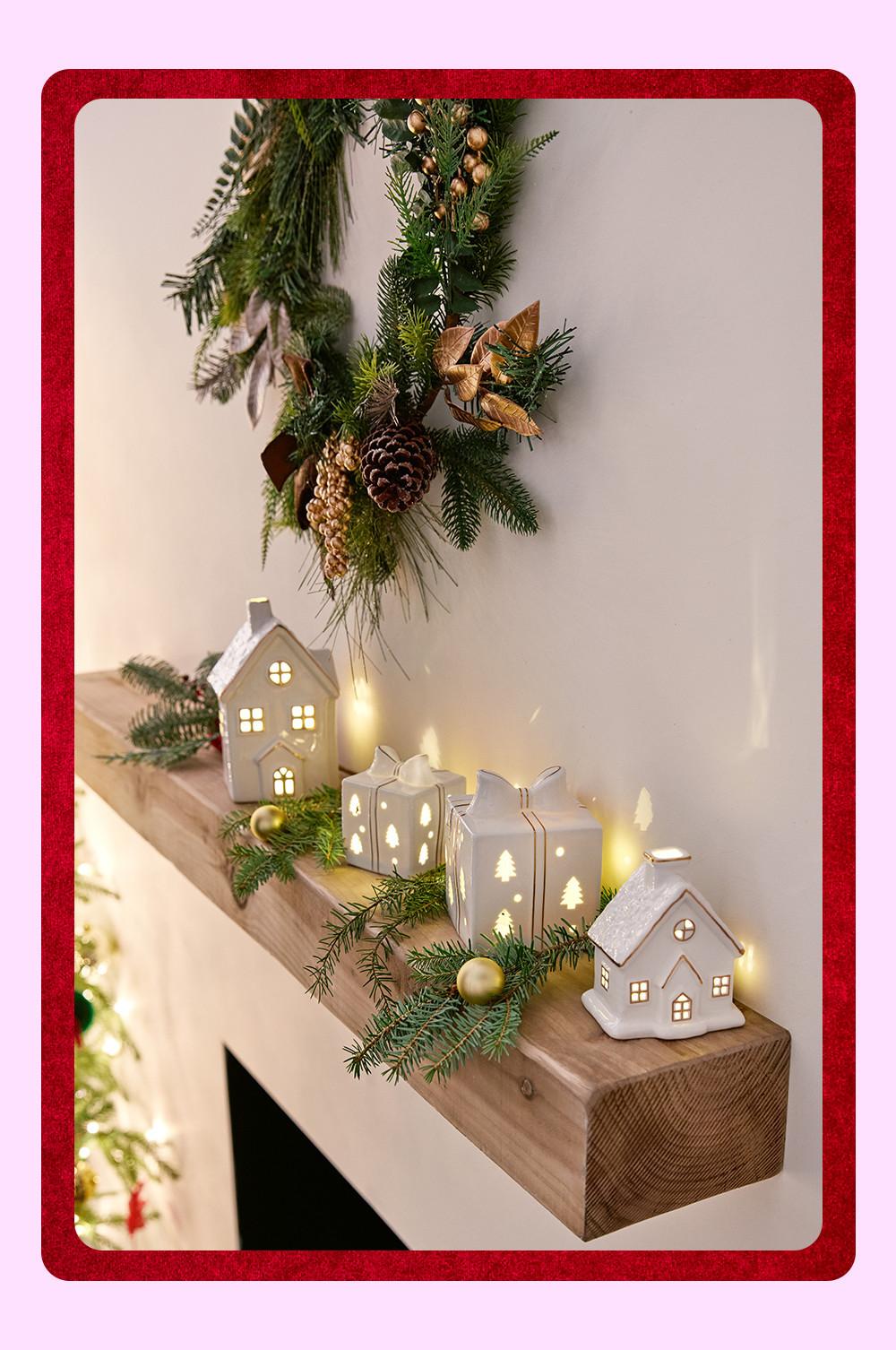 Repisa de chimenea decorada con casitas de cerámica iluminadas, cajas de regalo, ramas de pino, piñas y bolas de Navidad.