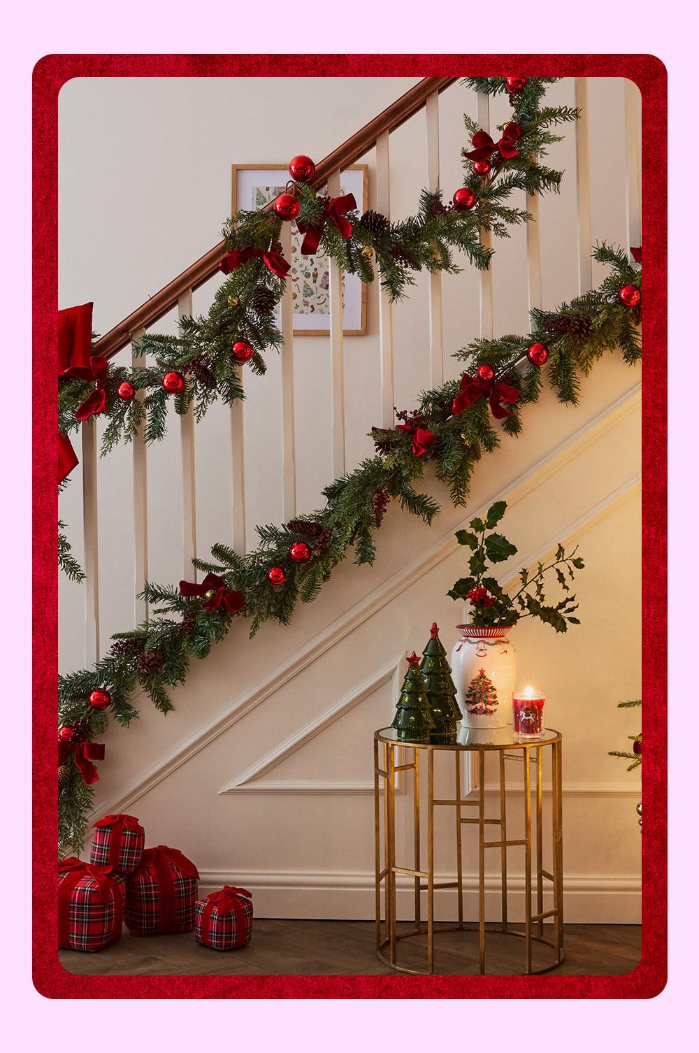 Escalera con decoración navideña compuesta por una guirnalda de hojas y ramas artificiales, bolas rojas y lazos, un árbol de Navidad y una mesilla auxiliar dorada.