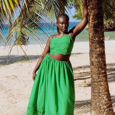 Model wearing a bright green two-piece outfit with lace crop top and flowing skirt on a tropical beach with palm trees and ocean view.