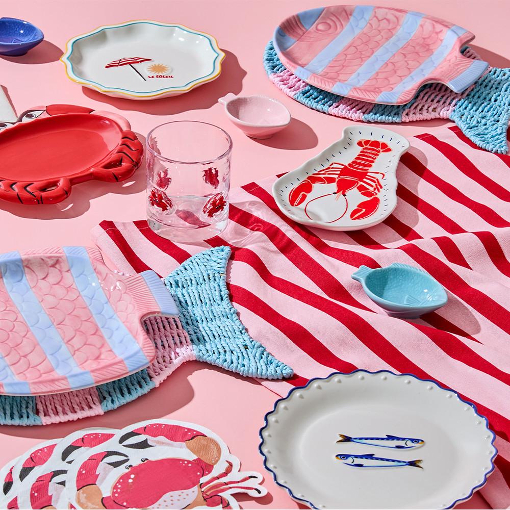 Piatti in ceramica con design di pesci e aragoste, piatto con granchio, bicchiere con stampe rosse, strofinaccio a righe.