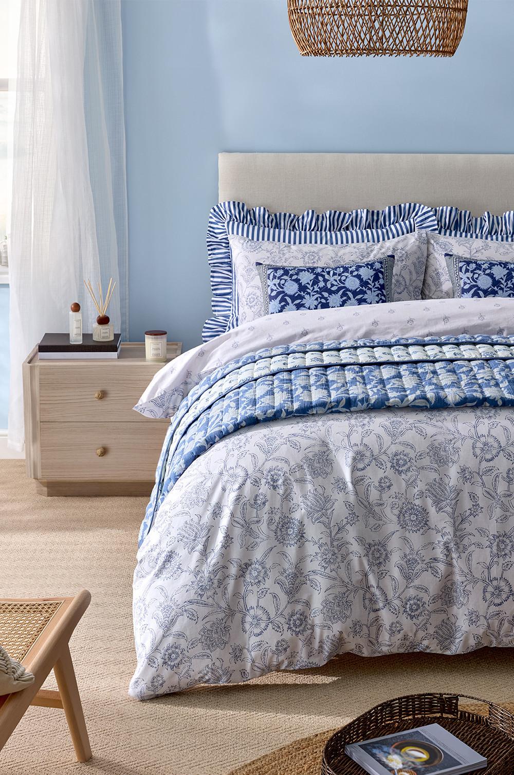 Blau-weiße Bettwäsche mit passenden Kissen mit Blumenmuster und gestreiften Rüschen, daneben ein Diffusor mit Stäbchen und eine Kerze.